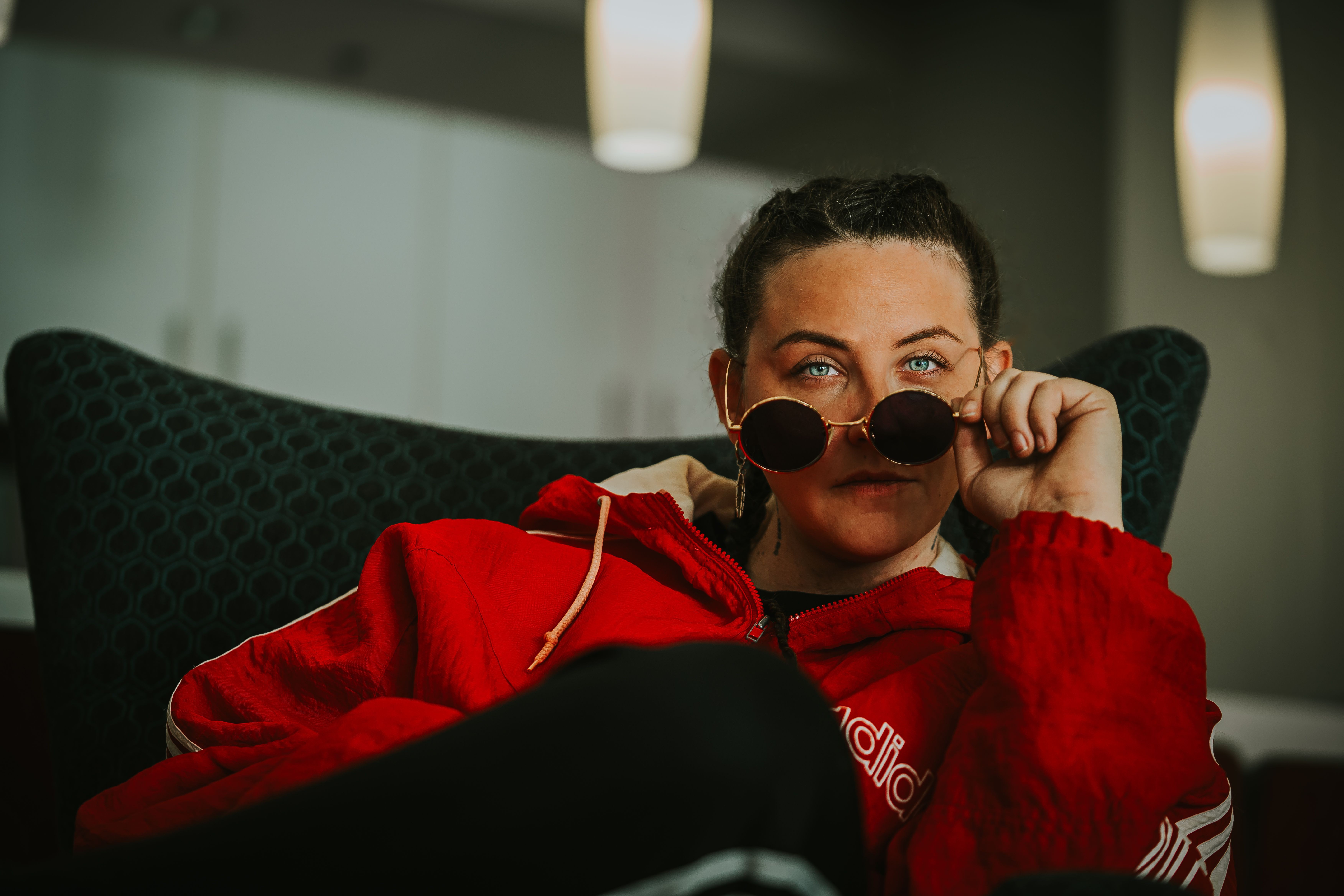Person with dark hair wearing sunglasses and a red hoodie, sitting on a dark chair in a dimly lit room.