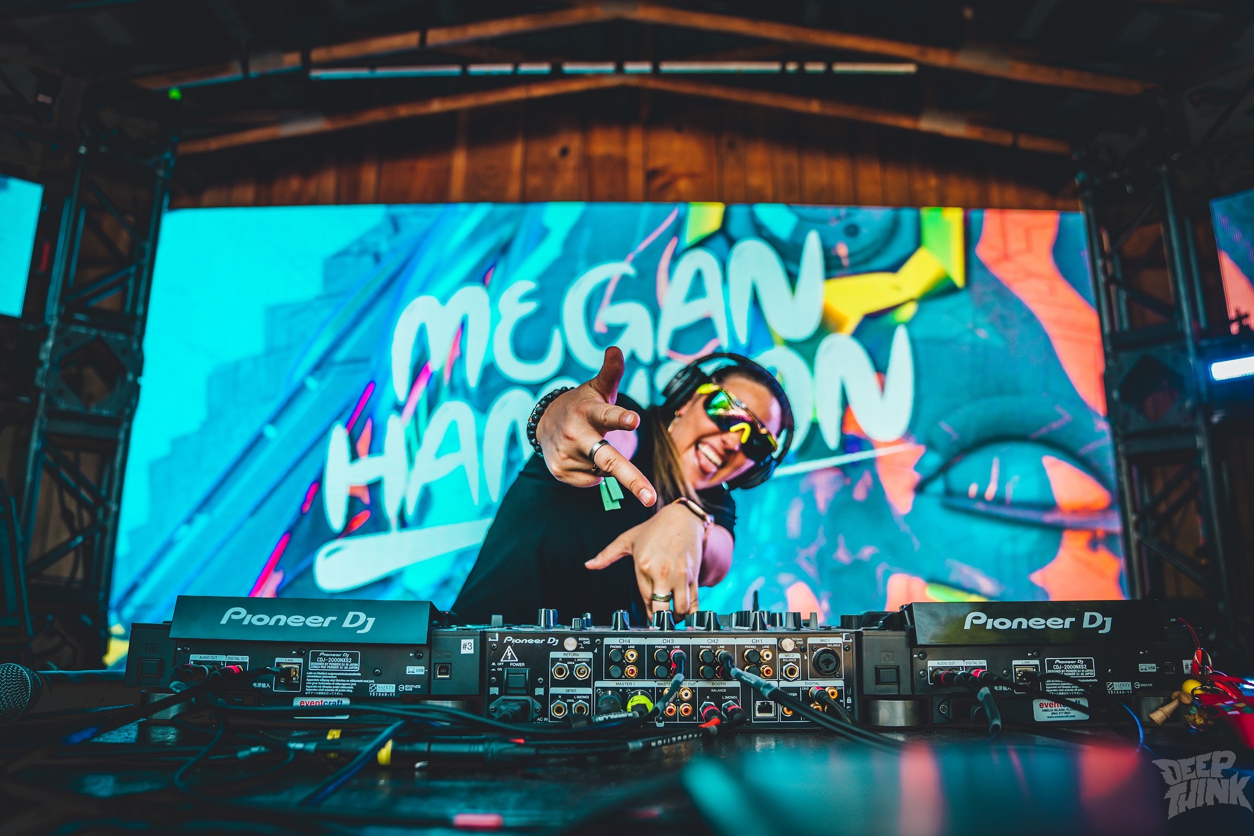 DJ wearing sunglasses and headphones, smiling and pointing at the camera, with a colorful digital background behind her.