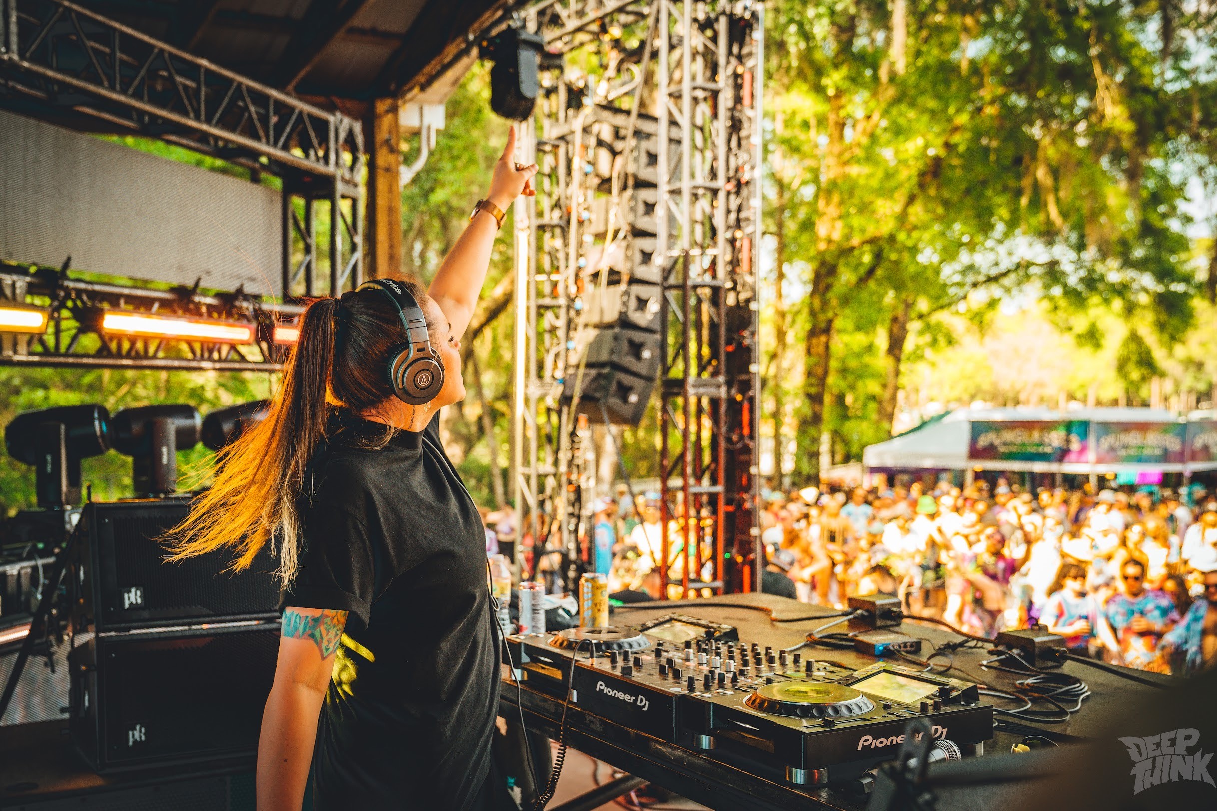 Female DJ wearing headphones performs on stage with a crowd outdoors in sunlight, trees in background.