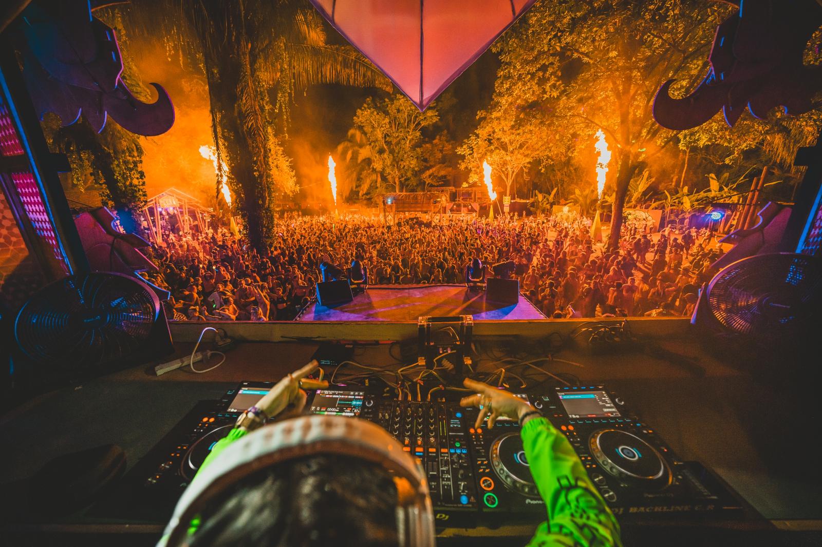 DJ performing at a concert with a large crowd and fiery stage in the background, viewed from behind the DJ booth.