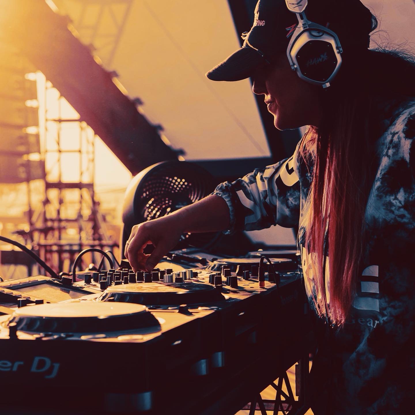 DJ wearing headphones operates equipment with sunset light in background.
