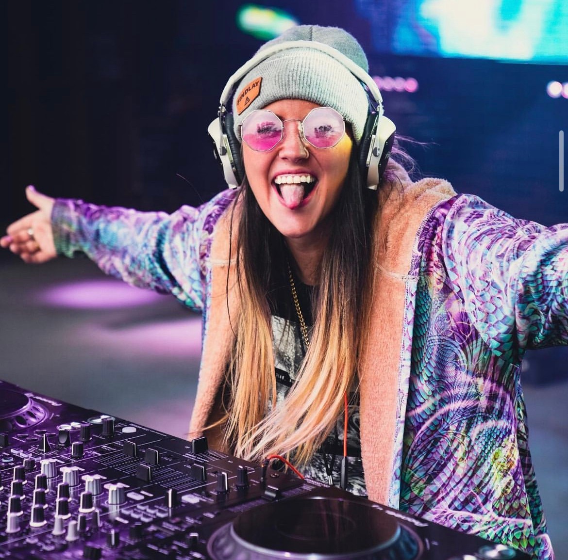 Female DJ wearing headphones, beanie, and sunglasses, smiling with arms outstretched behind DJ equipment in a colorful setting.