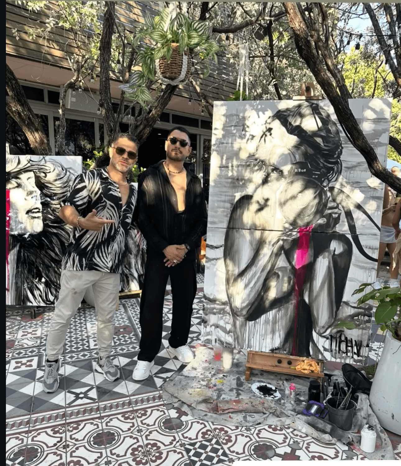Two people stand outdoors near a large painting of a woman with flowing hair, surrounded by trees and artwork, on patterned tile ground.