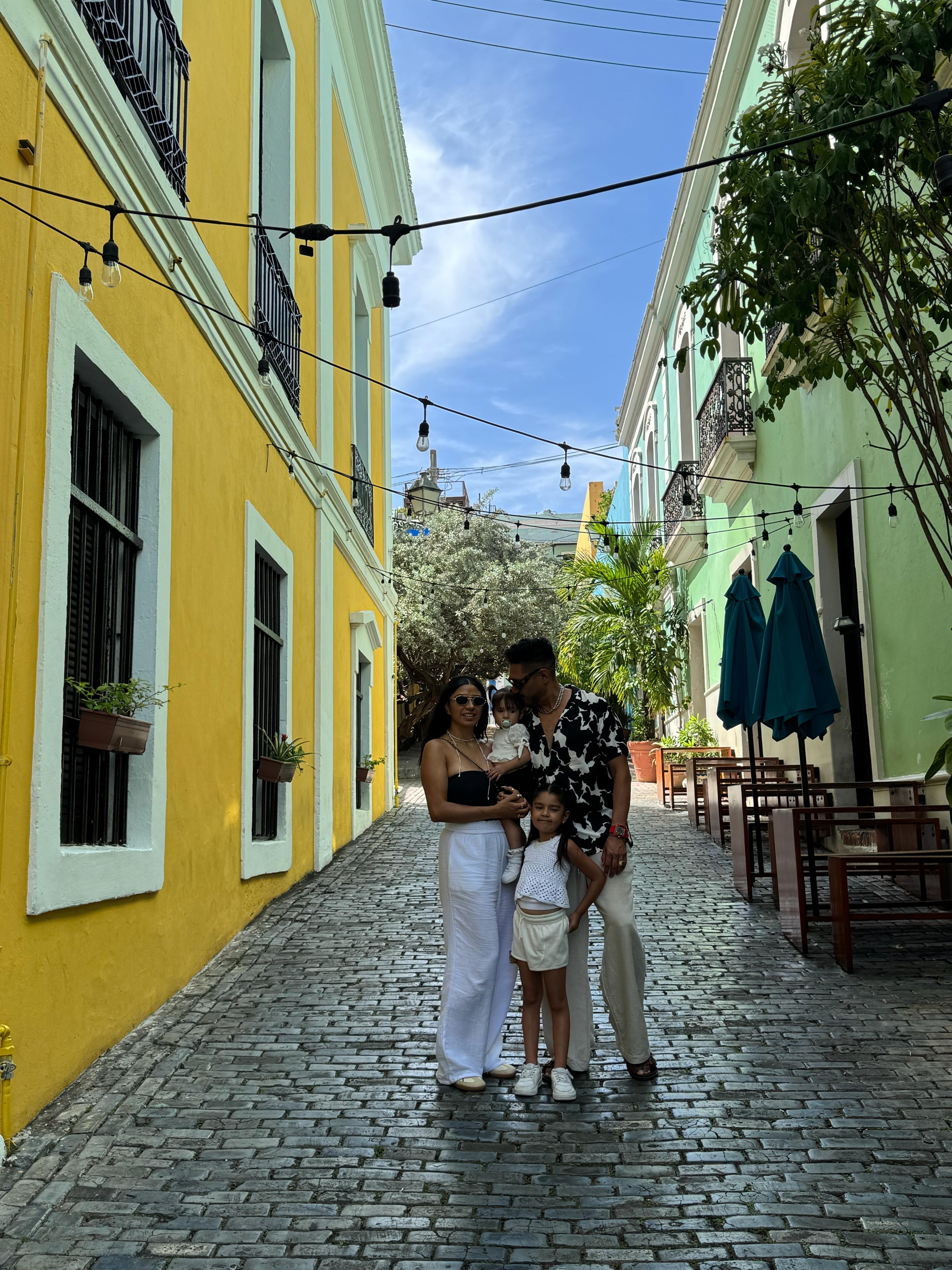 Family of three standing on cobblestone street between colorful buildings, with string lights overhead and a church in the background.