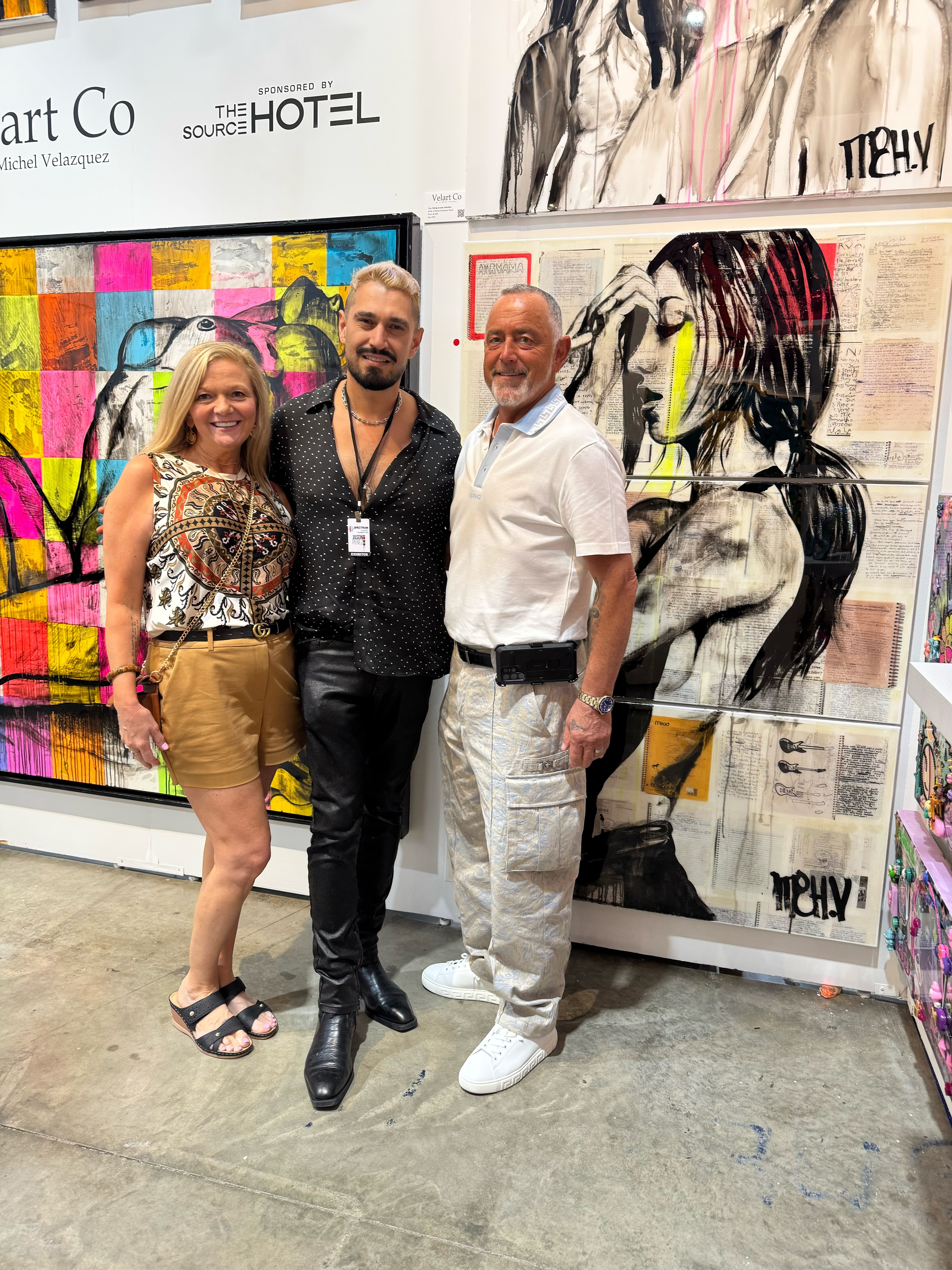 Three people standing in front of colorful artwork and a large black-and-white portrait at an art exhibition.