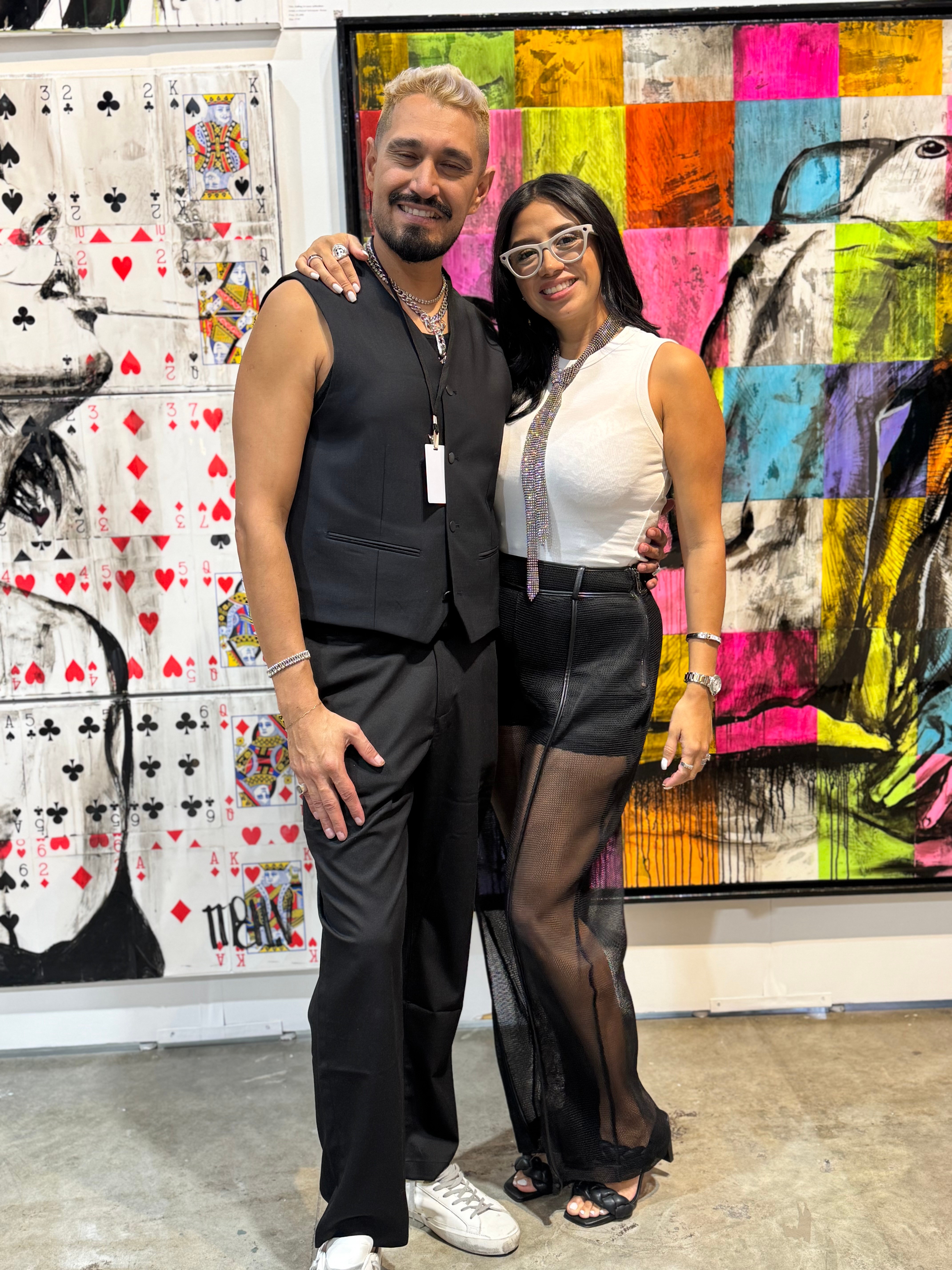 Two people standing together in front of colorful abstract art, smiling, with one person wearing a sleeveless top and the other in a white top and black skirt.