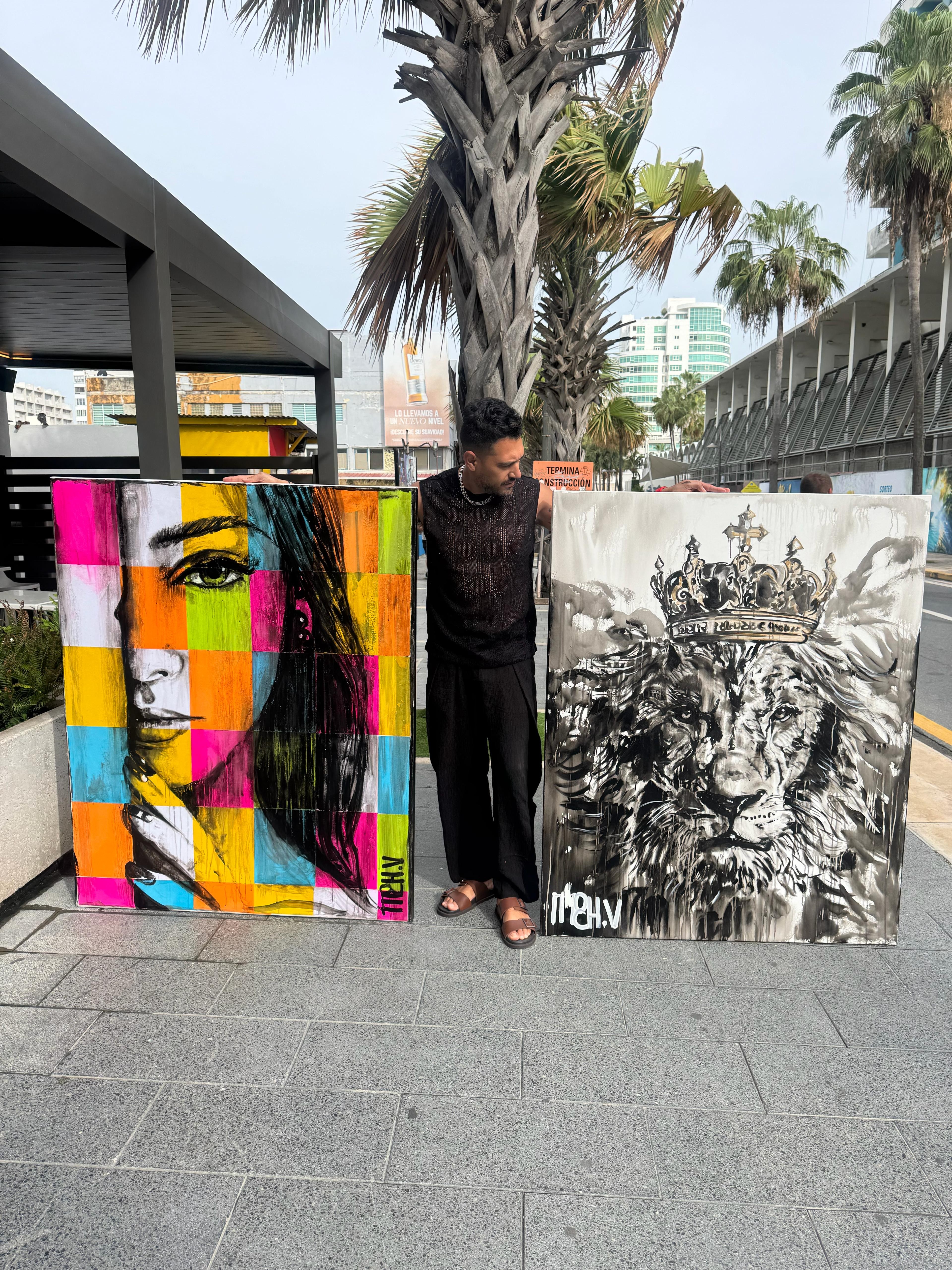 Person standing between two large paintings outdoors, one colorful abstract portrait and one black-and-white lion with a crown.