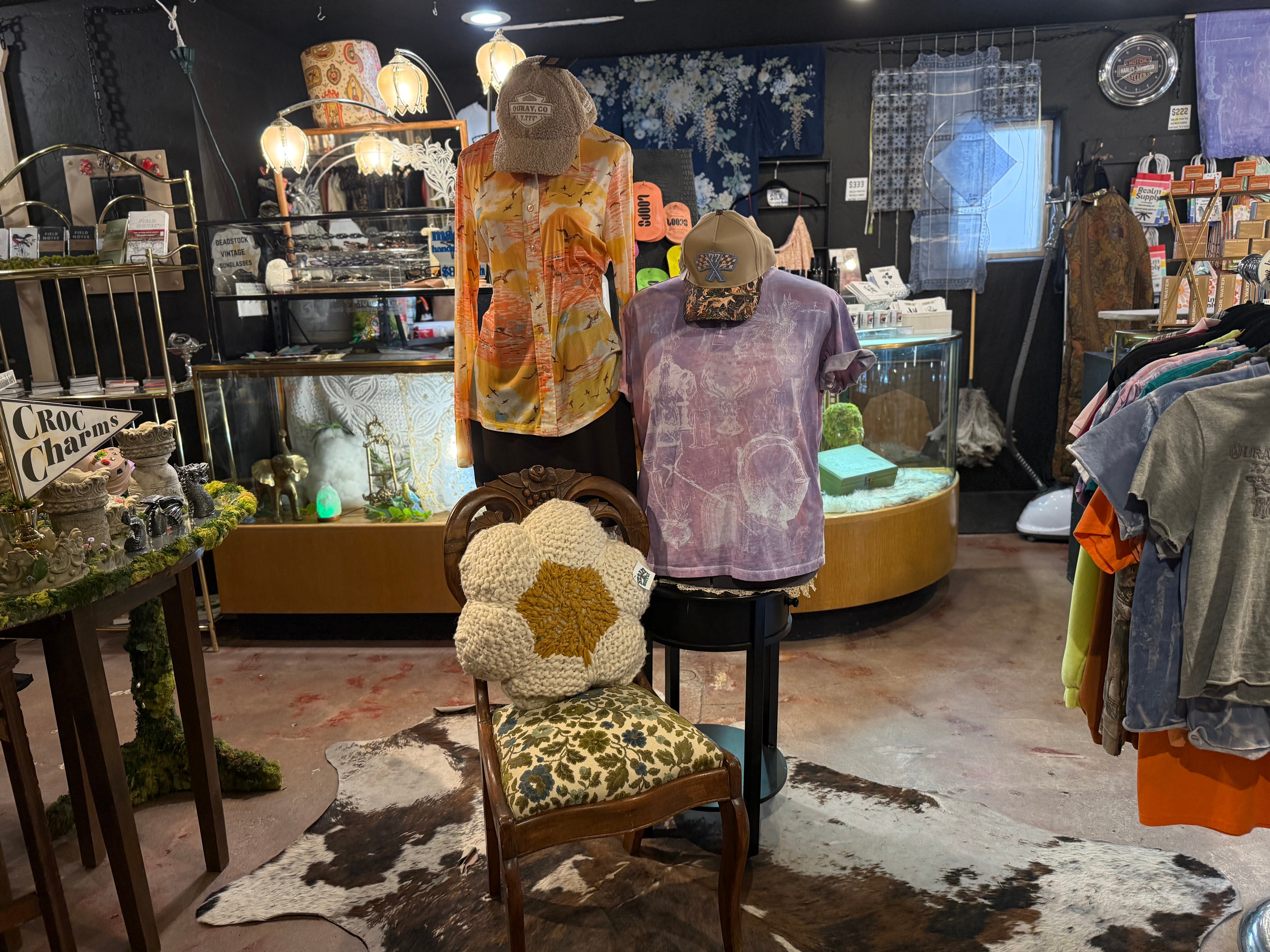 Display of mannequins wearing colorful tops, with a chair in front, inside a store with various items and clothing.