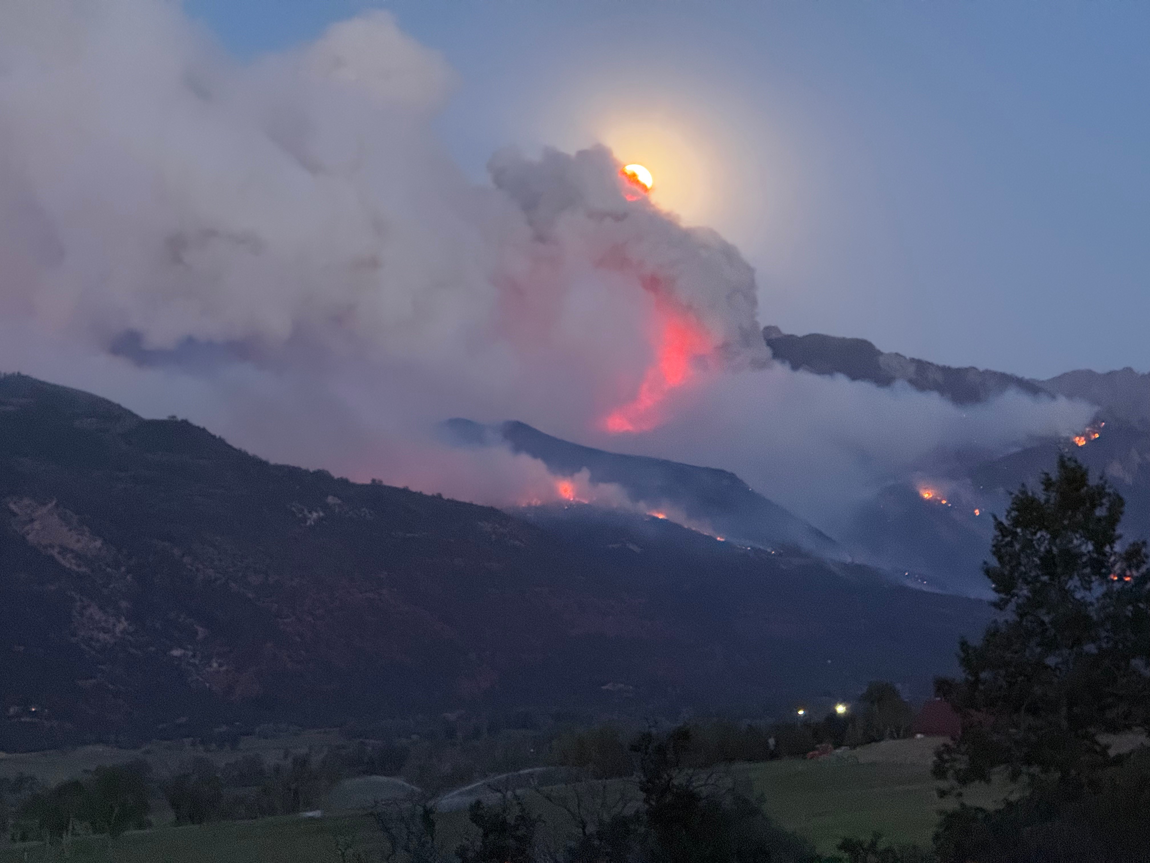 Volcanic eruption with smoke and ash rising over mountains, with the moon visible in the sky, and trees in the foreground.