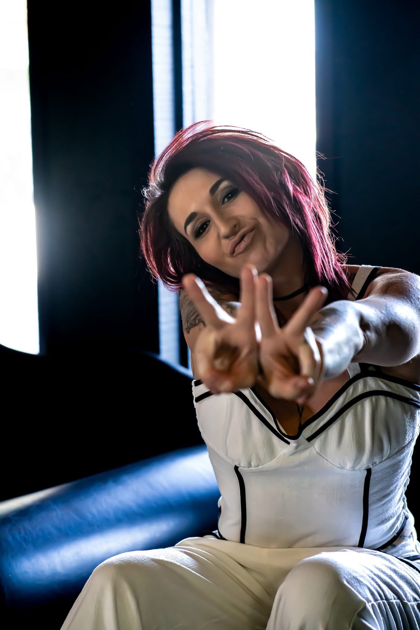 Woman with short red hair smiling and making a peace sign with her hand, sitting indoors near a window.
