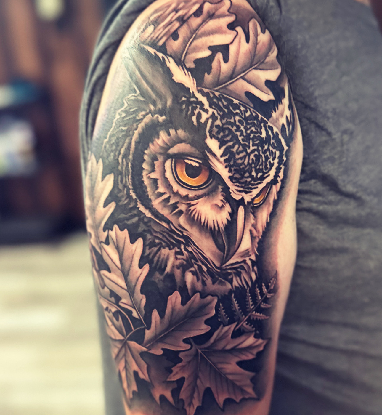 Tattoo of an owl with detailed feathers and leaves on upper arm, in black and gray shading.