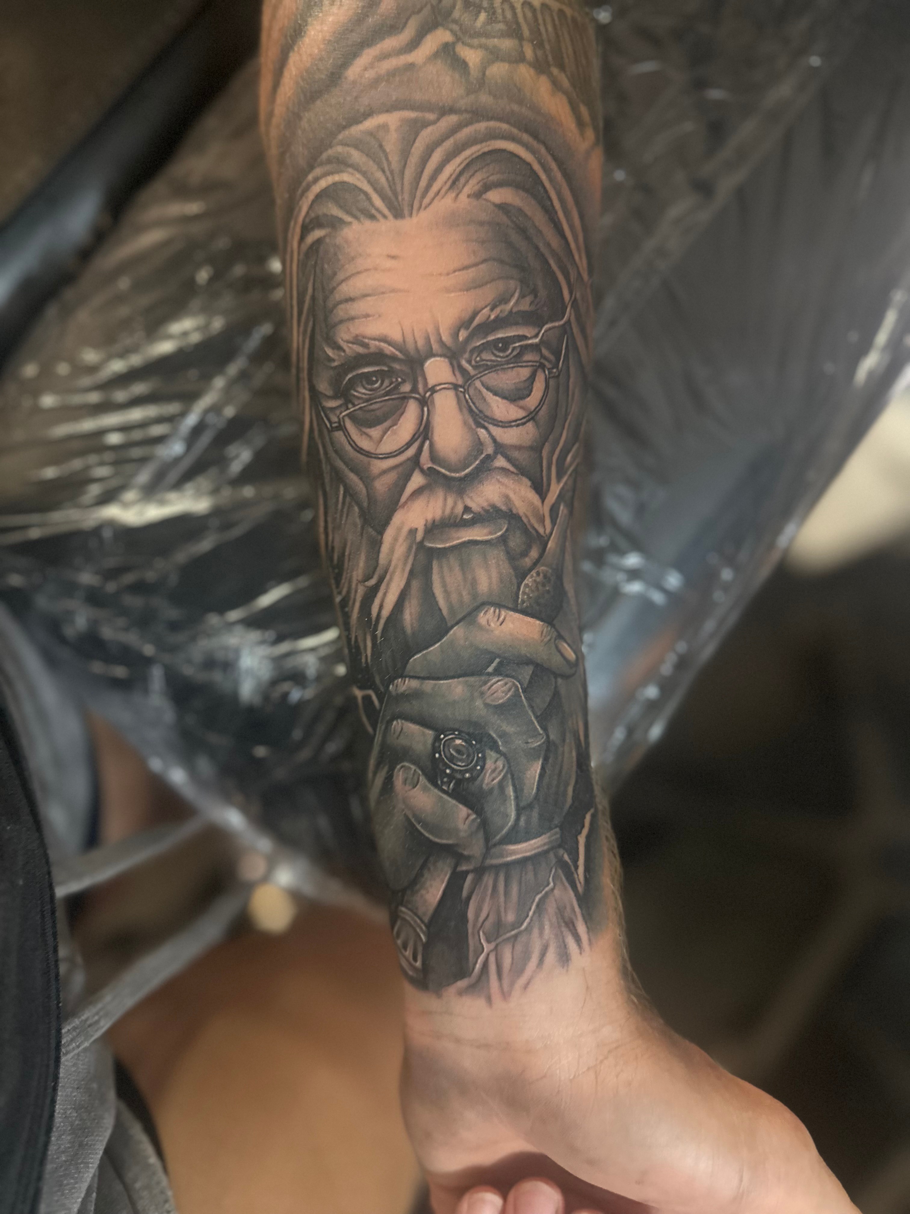 Tattoo of an elderly man with glasses, beard, and thoughtful expression on forearm.