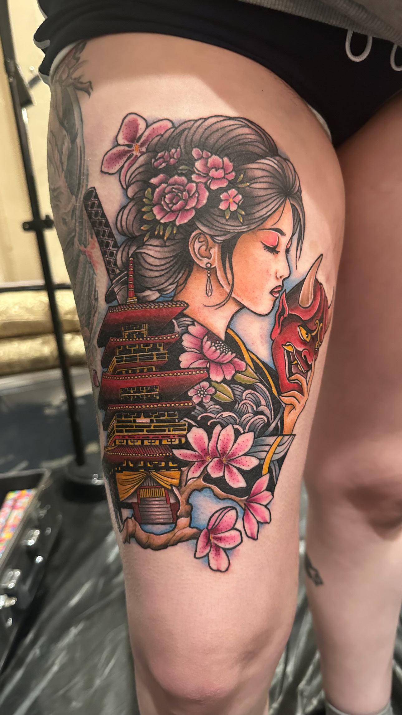 Tattoo of a woman with flowers in her hair, holding a sword, surrounded by pink blossoms, on thigh.