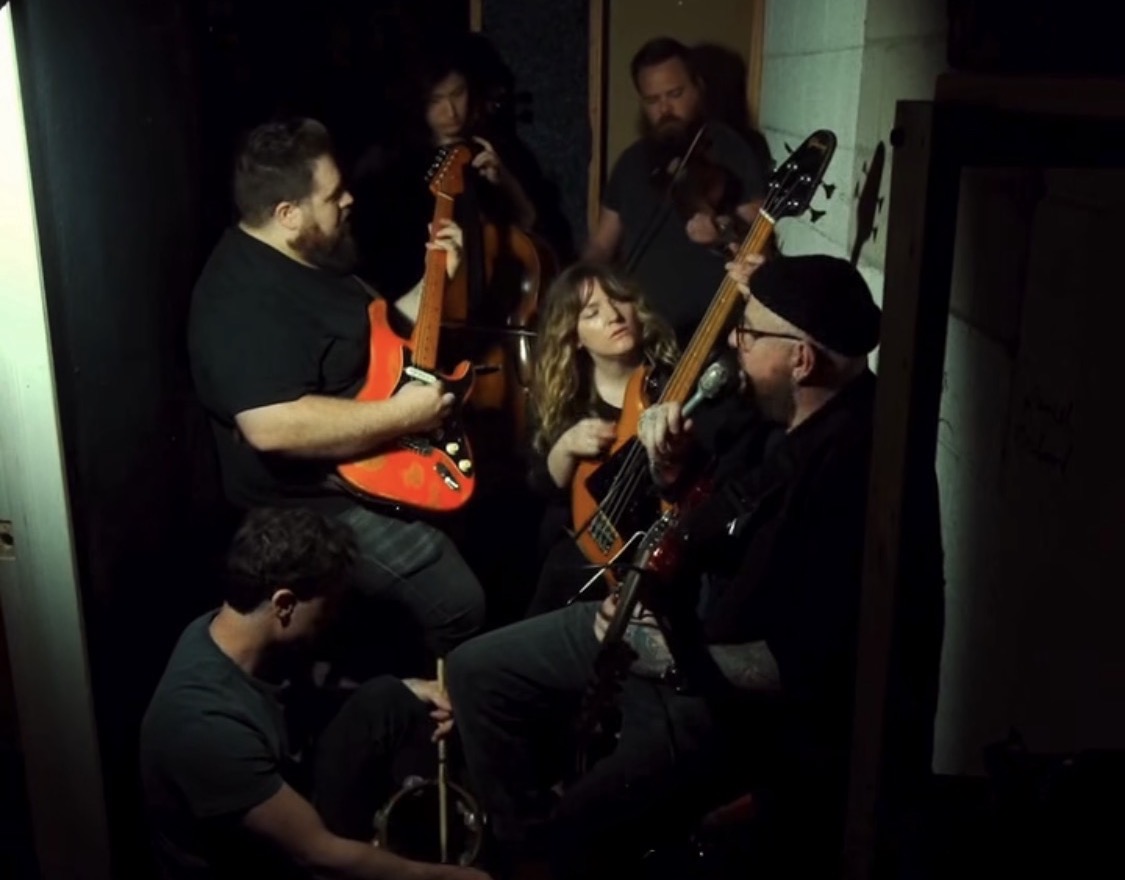 Group of five musicians playing guitars and a ukulele in a small, dimly lit room.