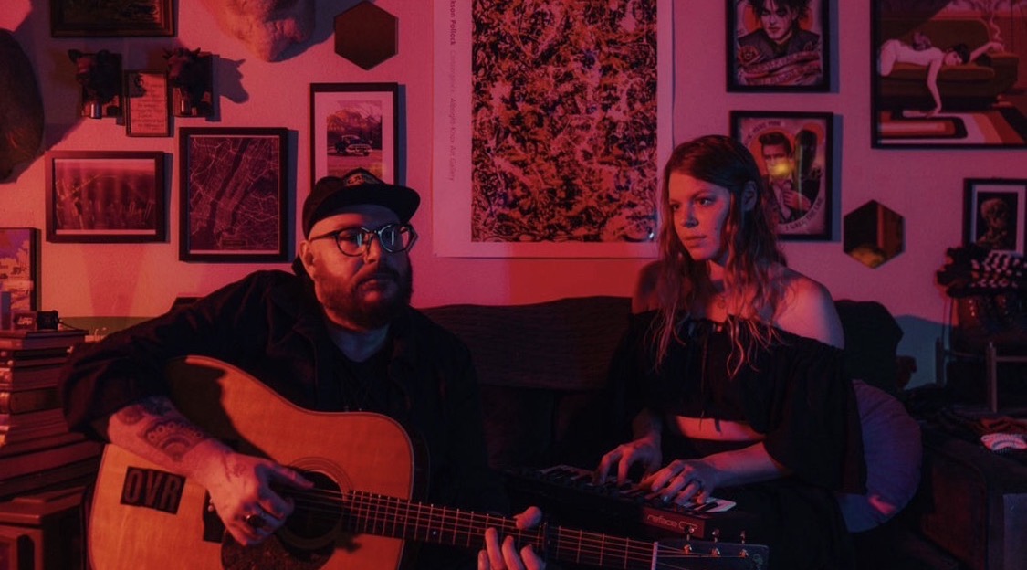 Two people sit on a couch in a room with framed pictures, one playing guitar, the other listening, illuminated by pink and purple lighting.
