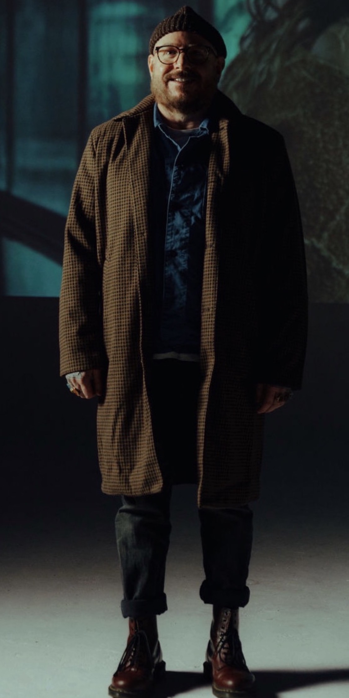Man standing in a dimly lit environment, wearing a coat, glasses, and boots, with a dark background.