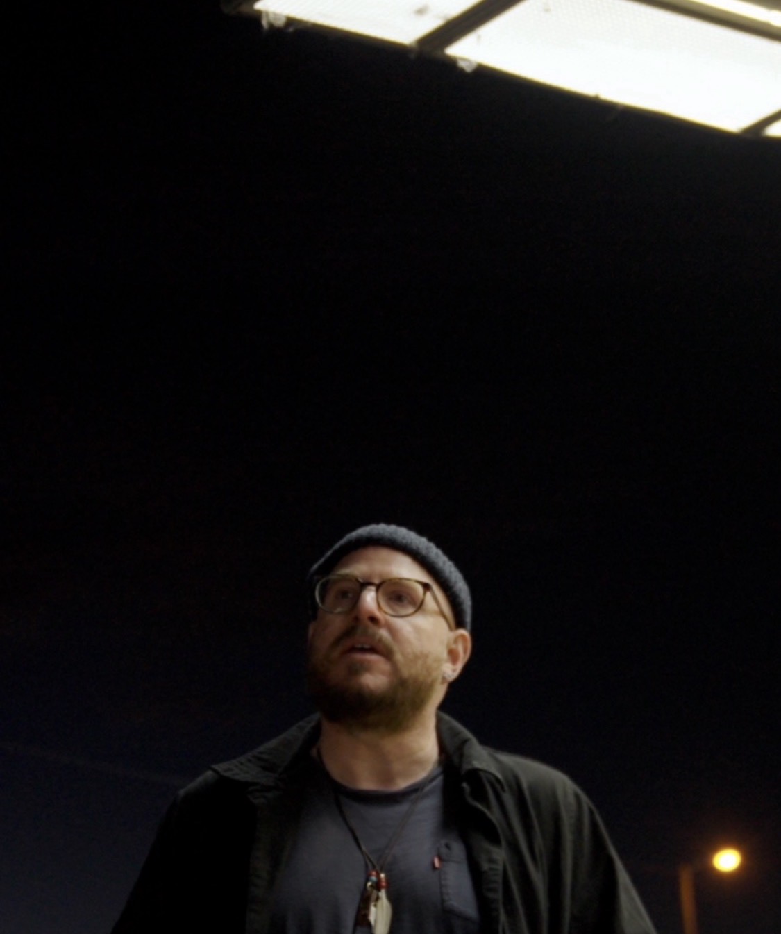 Man with glasses and a beanie, wearing a dark jacket, looking upward, against a dark background with a light source above.