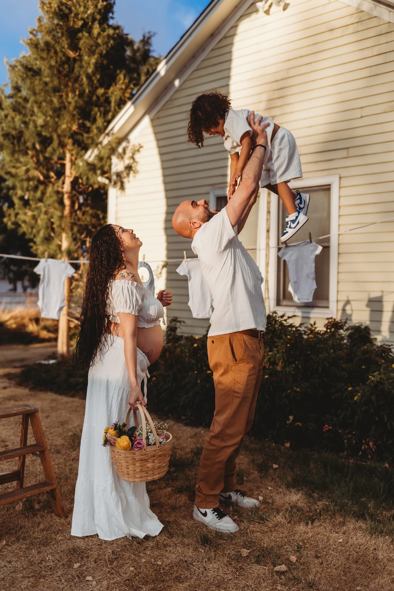 A man lifts a child in the air while two women watch outside a house with a window and trees in the background.