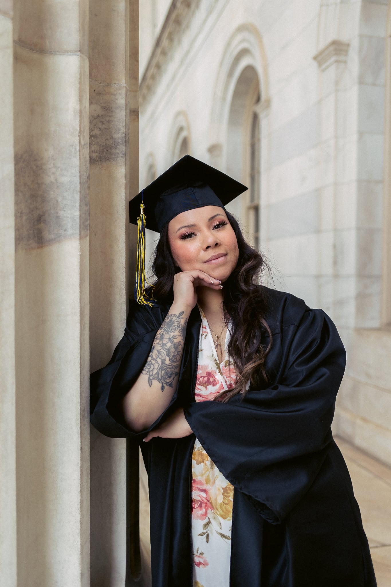 Young woman in graduation gown and cap leaning against a stone column inside a building with arches.