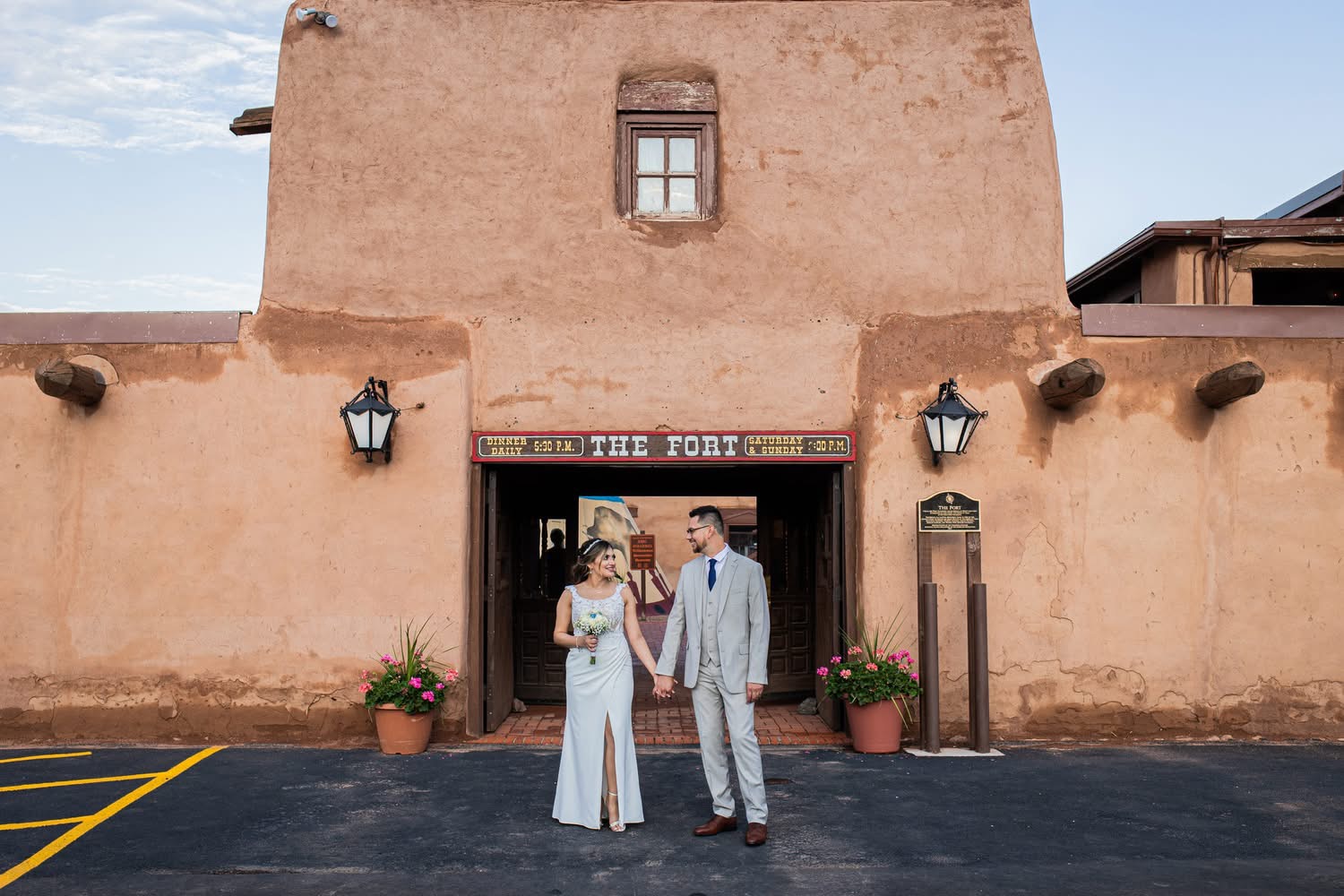 A couple stands in front of a building with a sign that reads 'The Fort,' holding hands and looking at each other.