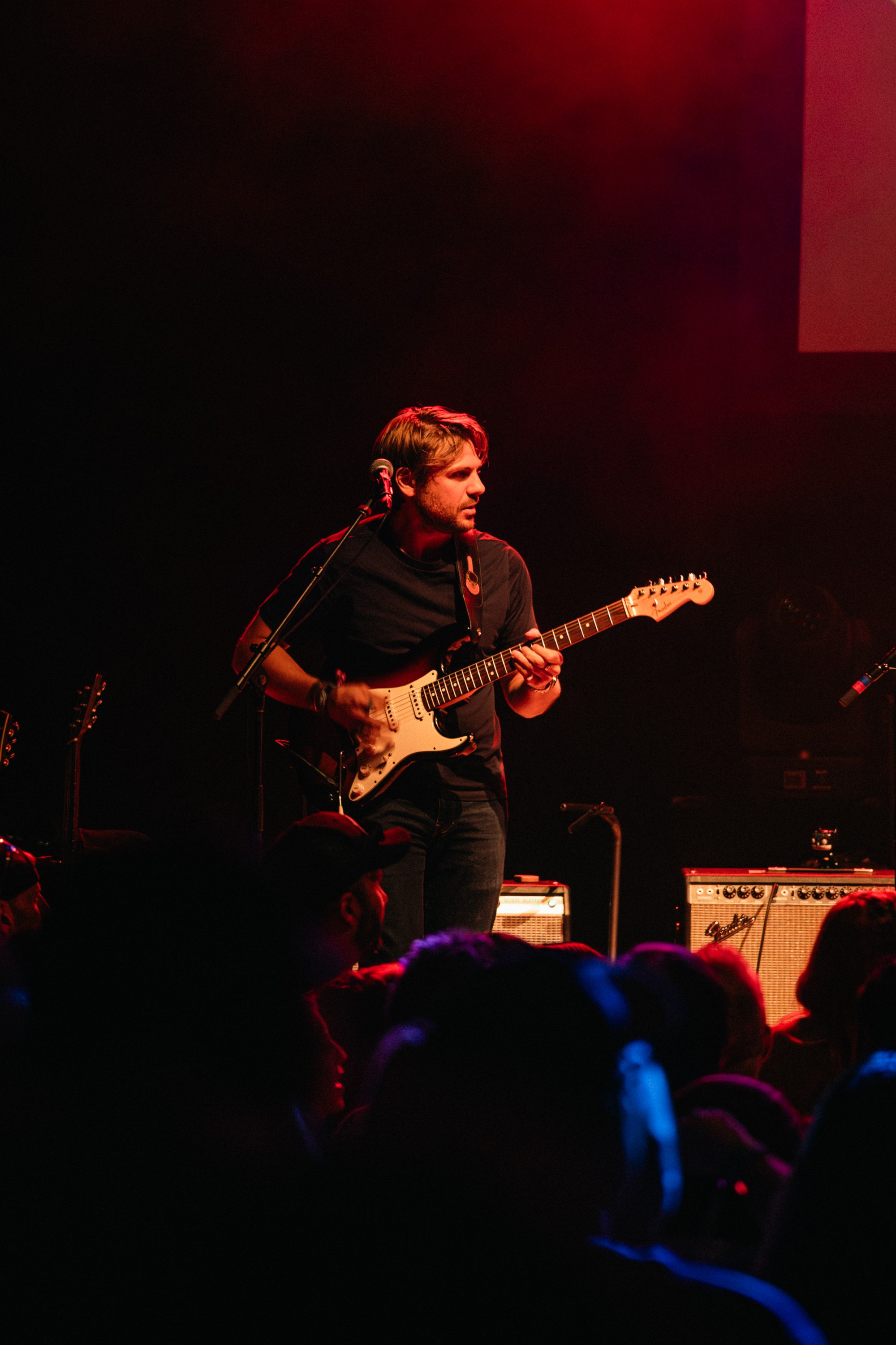 Musician playing an electric guitar on stage with red lighting, audience in foreground, dark background.