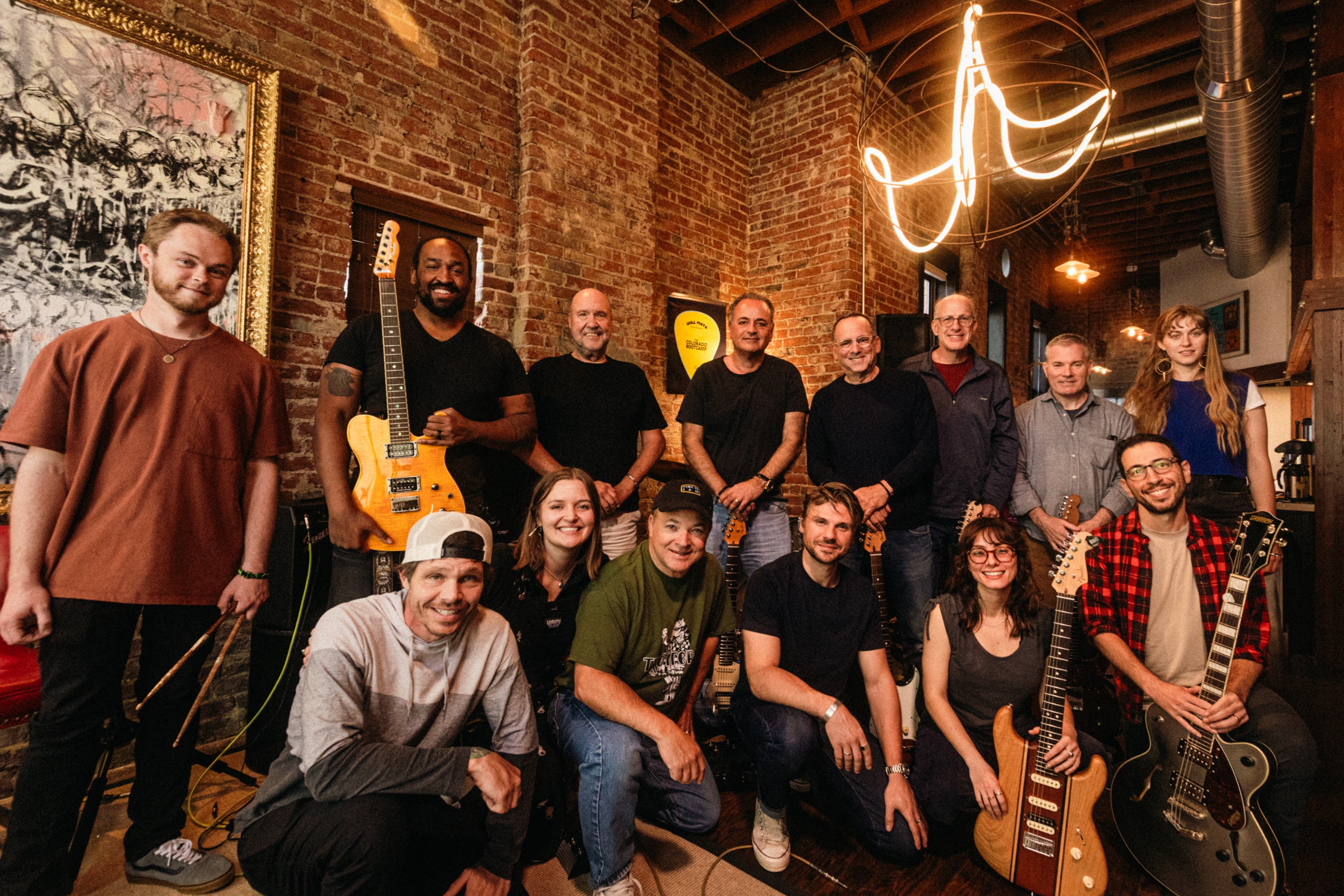 Group of 13 people in a music studio with guitars and artwork, smiling and posing for a photo.