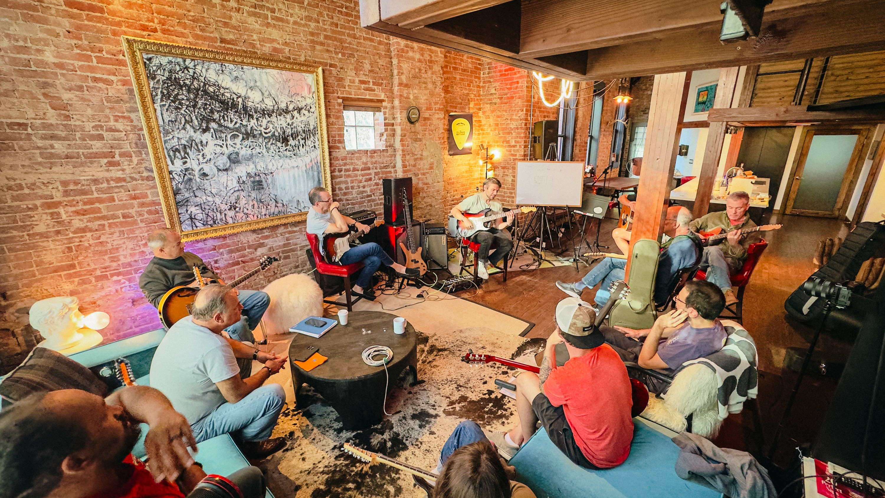 Group of people sitting in a cozy room watching a live music performance with guitars and microphones, brick walls, and artwork.