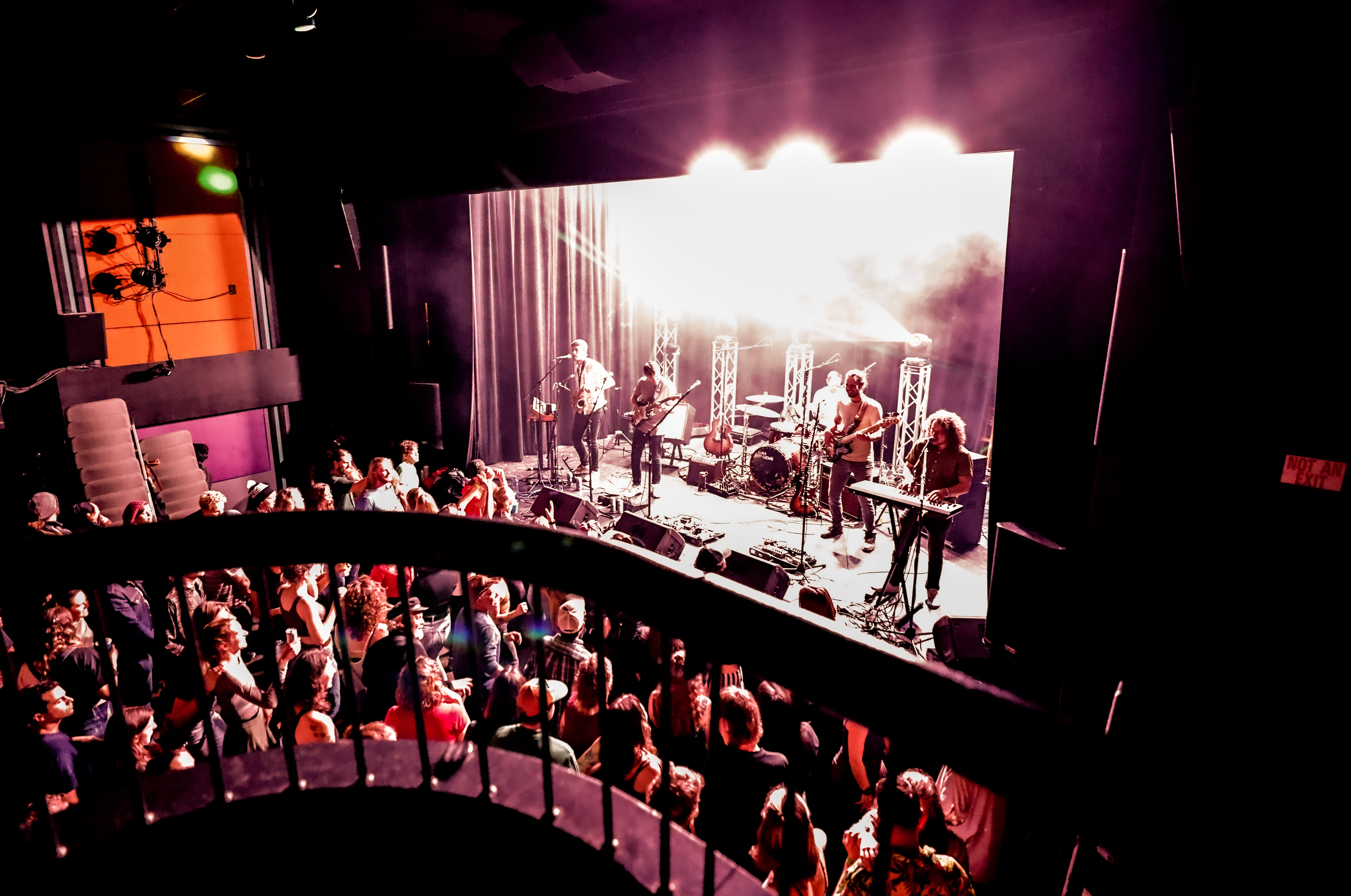 Band performs on stage with bright lights, audience watching in a dark venue, balcony railing visible in foreground.
