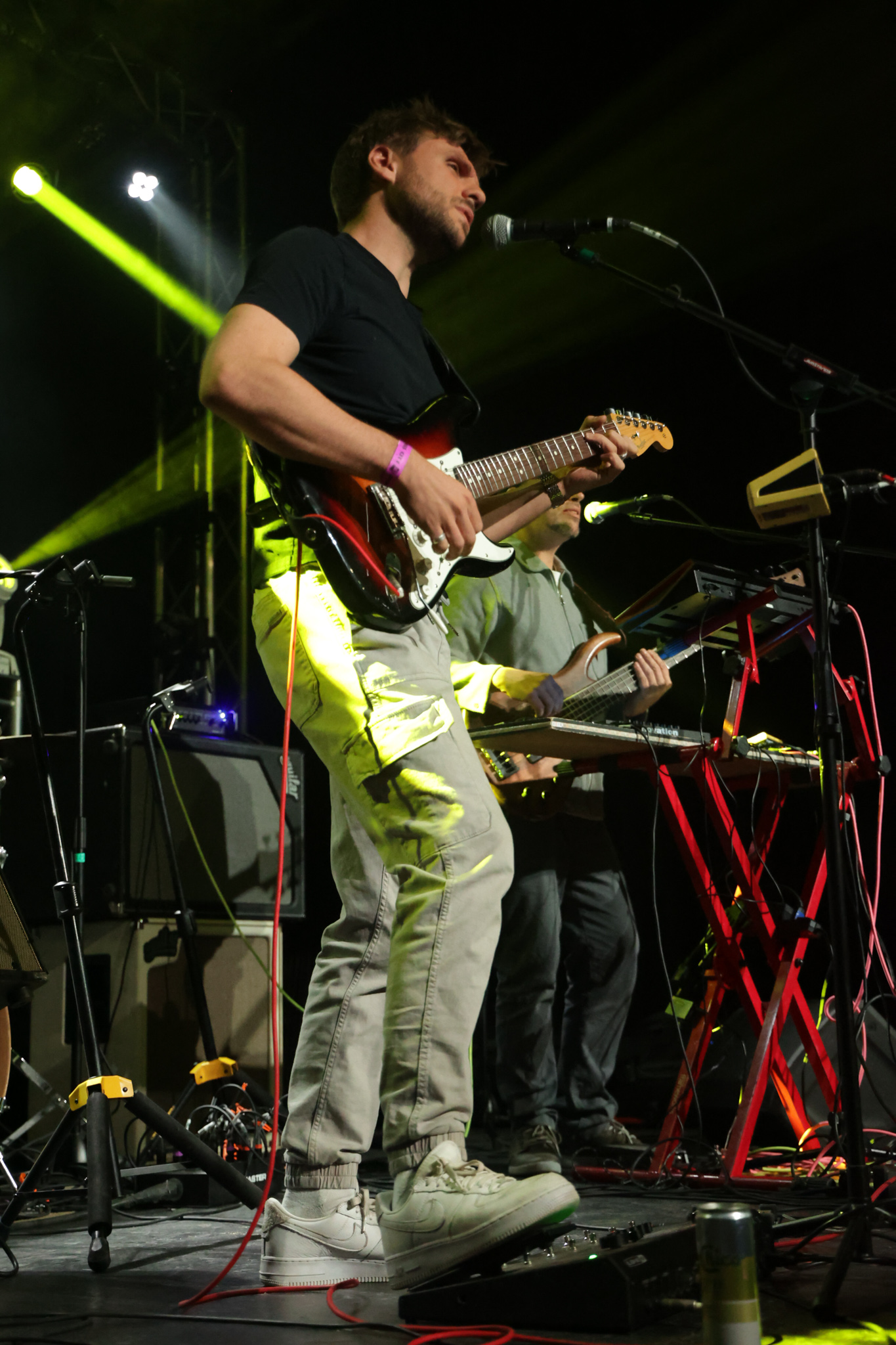 Musician playing guitar and singing into microphone on stage with keyboard and equipment, yellow stage lights, dark background.