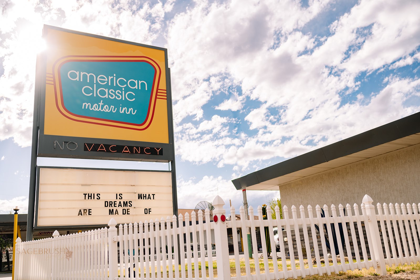 Sign for American Classic Motel Inn with a partly cloudy sky in the background, white picket fence in front, and a building to the right.