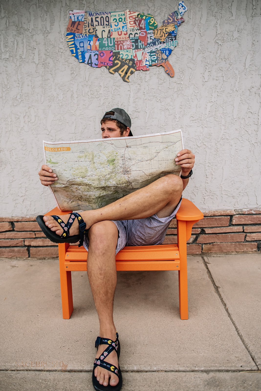 Person sitting on orange bench reading a large map, with a map of the United States made of license plates on the wall behind.