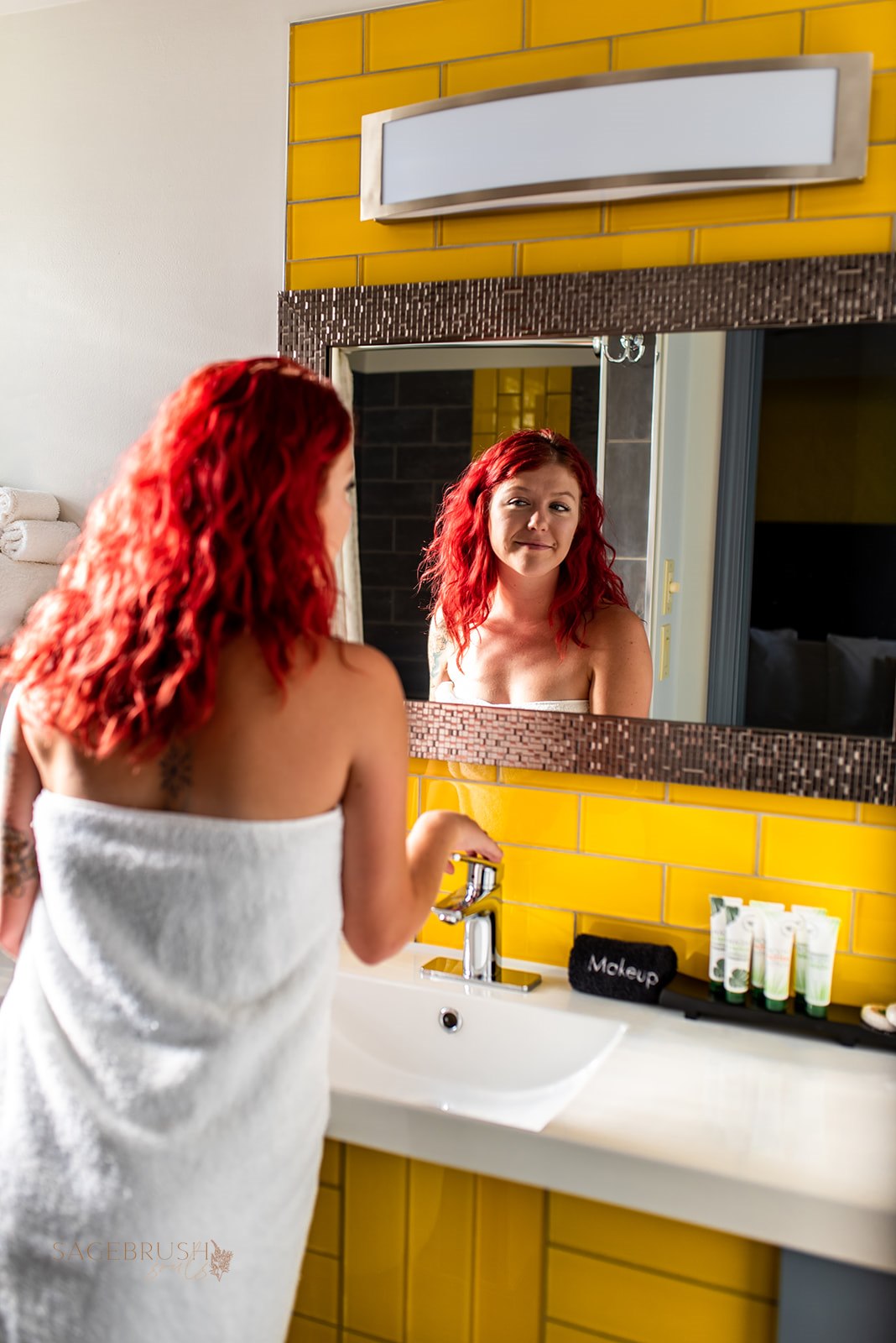 Woman with red hair looking at her reflection in a mirror above a bathroom sink, yellow tiled wall behind her.