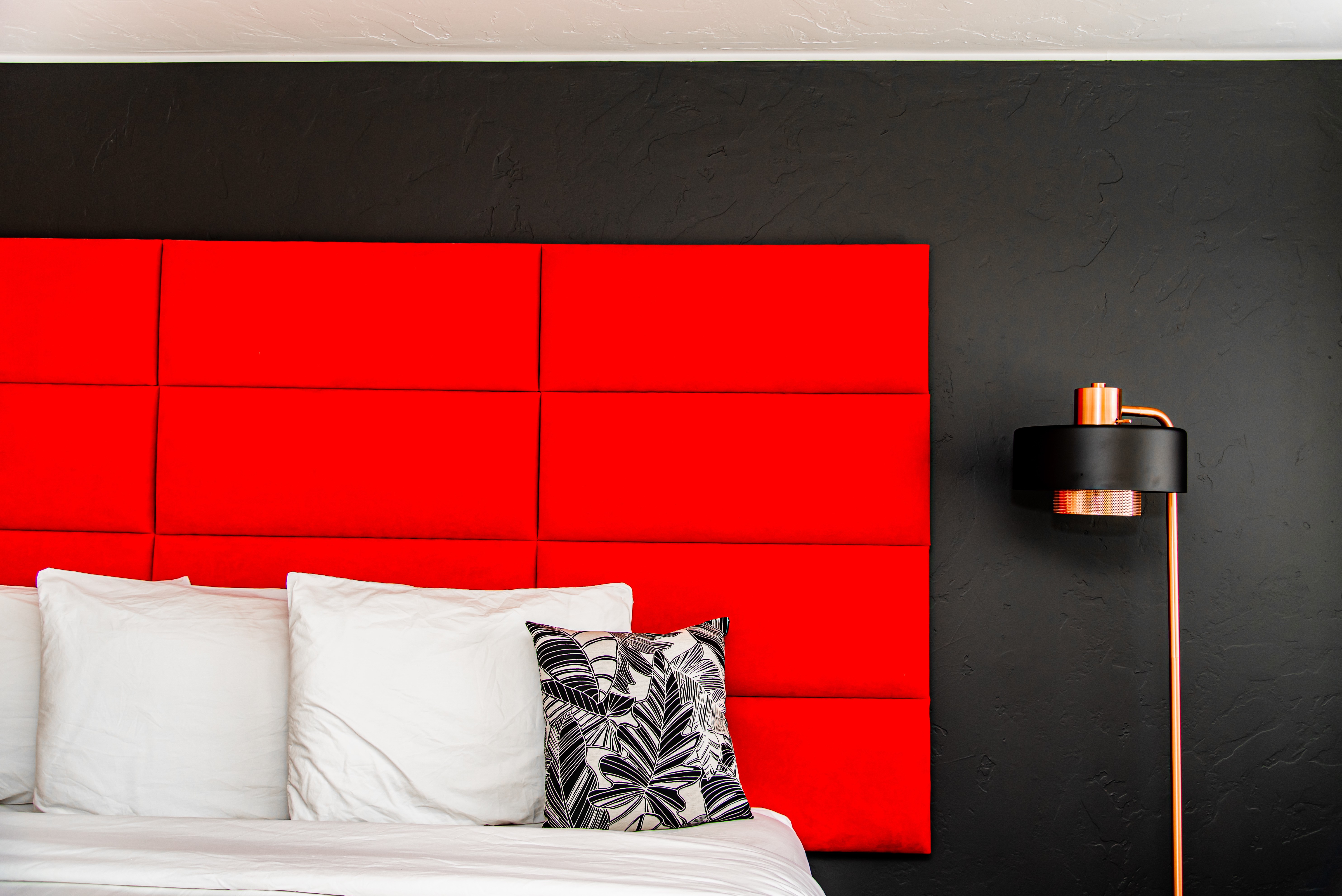 Red headboard with white pillows and a black and white patterned pillow, black wall, and a black and copper floor lamp.