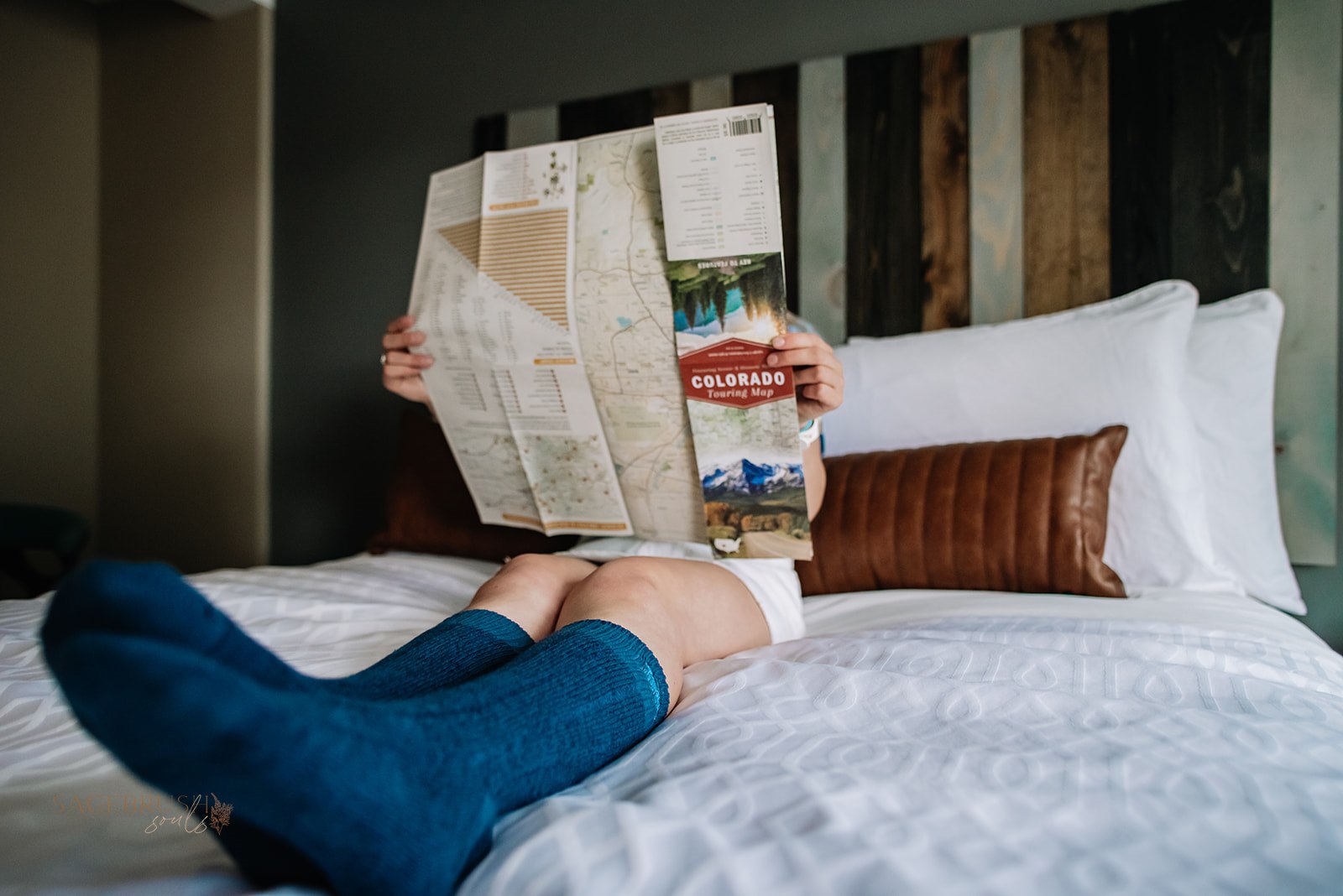 Child sitting on bed reading a newspaper, legs stretched out, wearing blue socks and shorts, head obscured by newspaper.