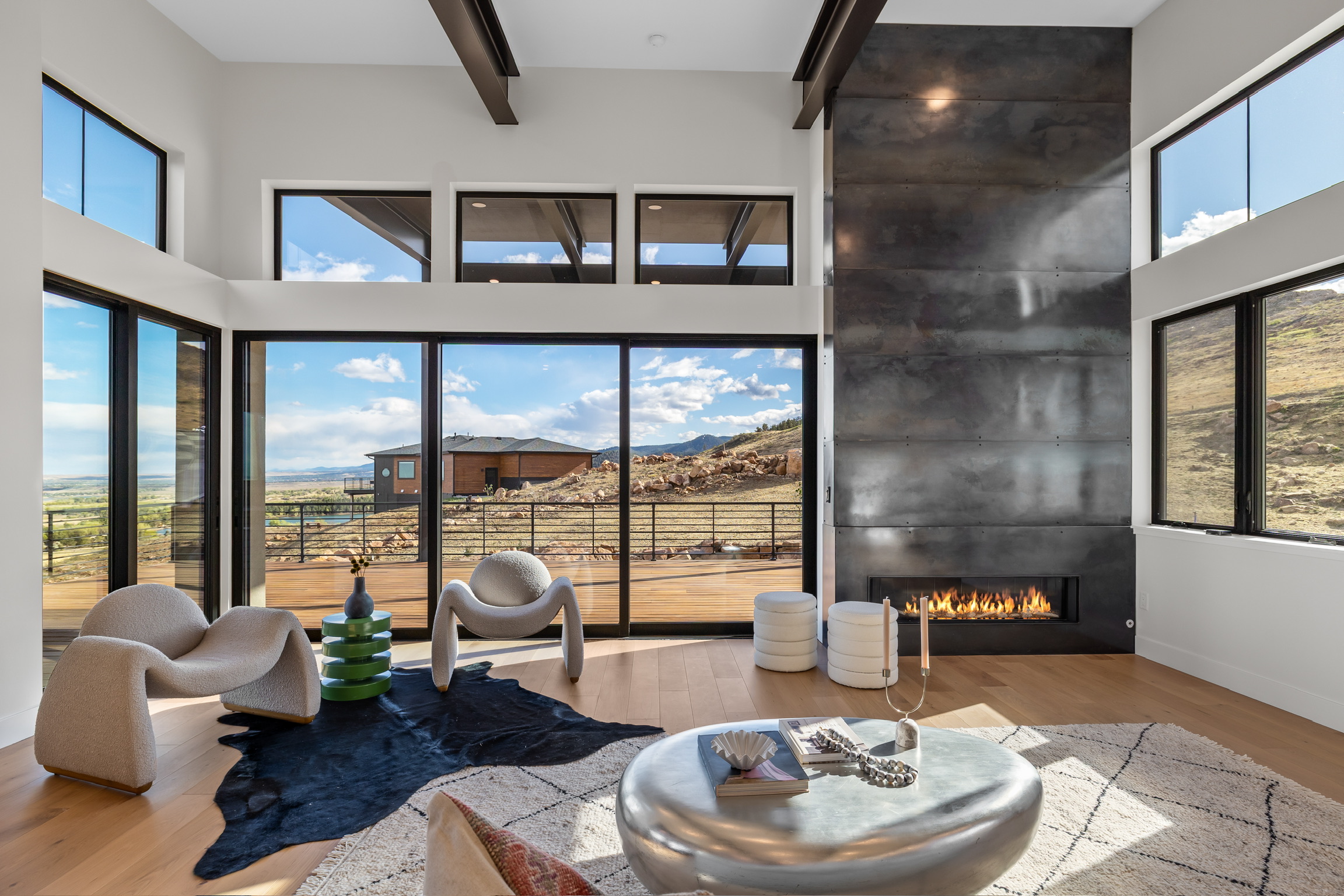 Living room with large windows, modern fireplace, and seating area with chairs and a rug, overlooking outdoor landscape.