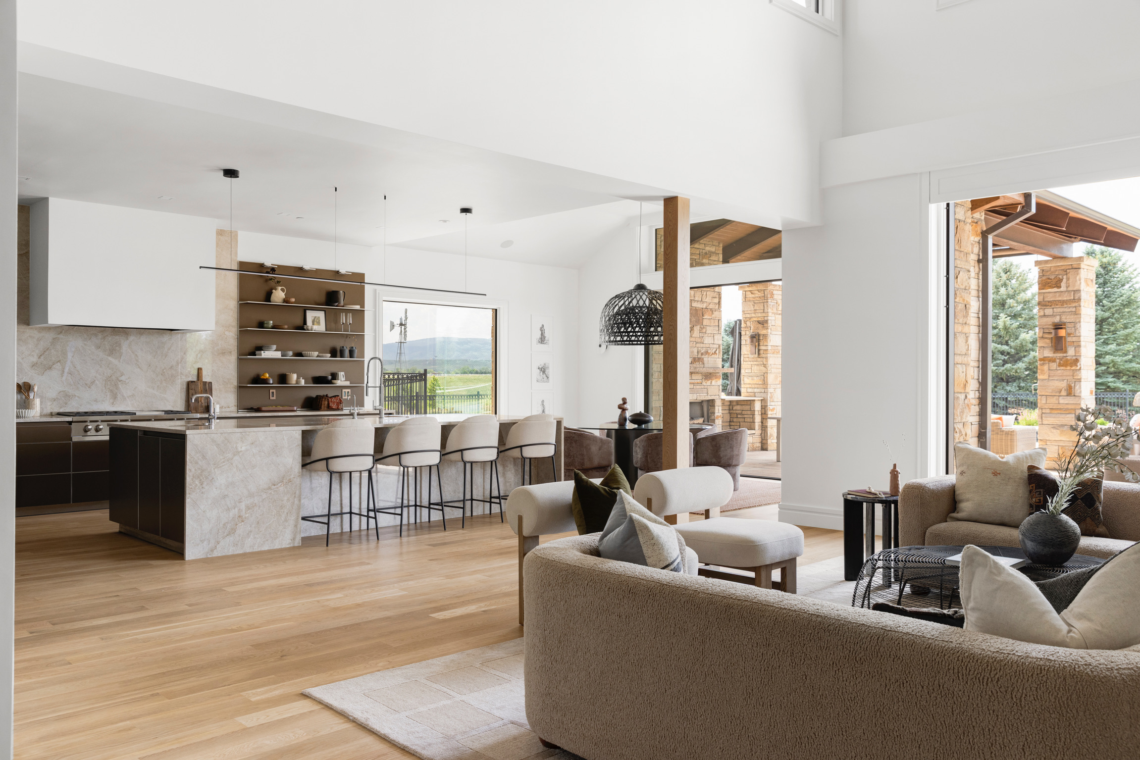 Open-concept living area with kitchen, bar stools, and seating, bright with large windows and wooden accents.