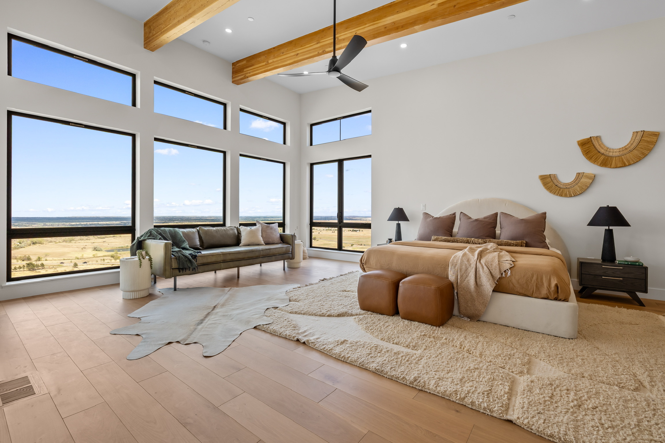 Bedroom with large windows, bed with pillows, nightstands, and a sitting area with a sofa, rug, and decorative wall art.