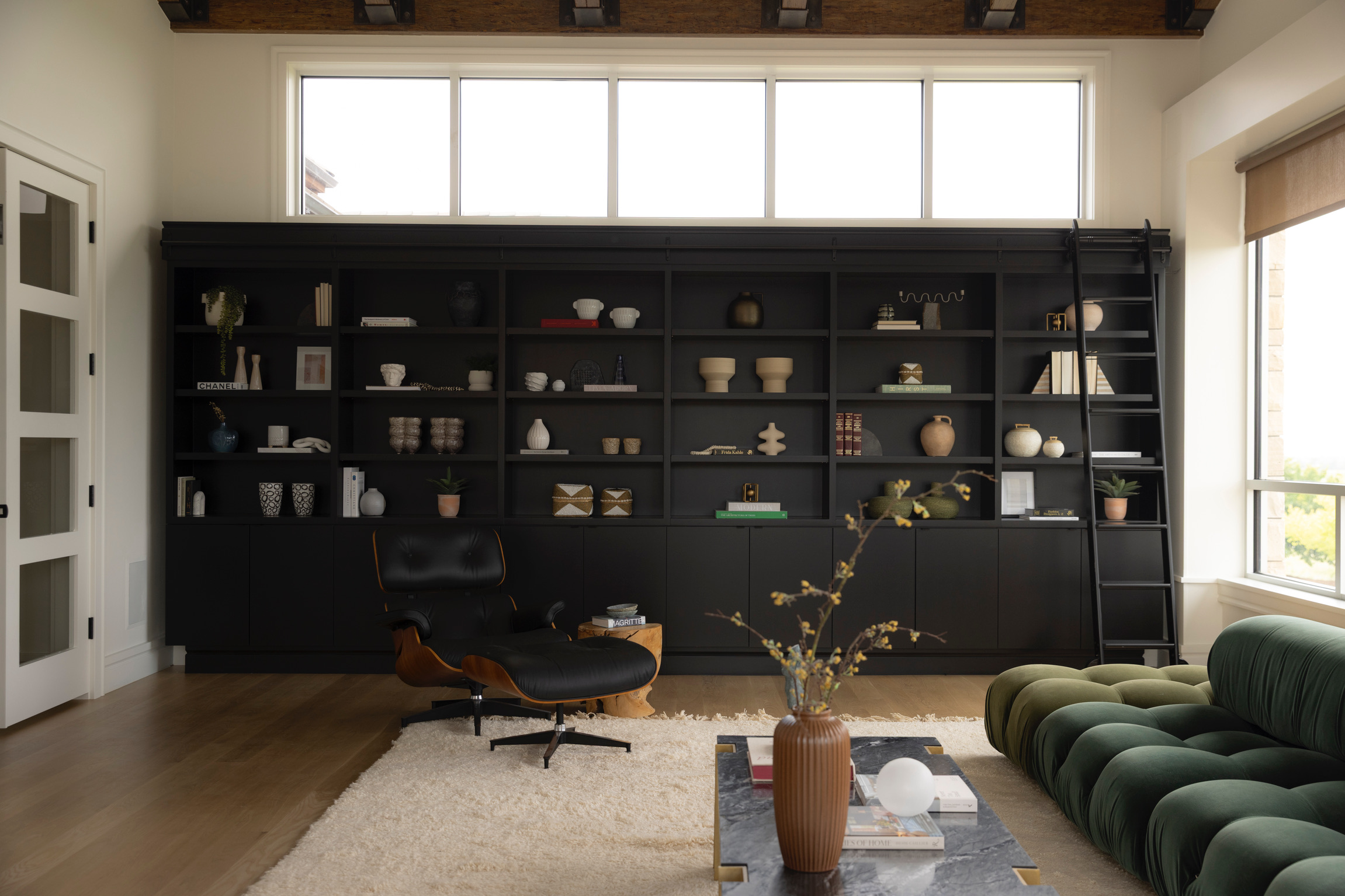 Living room with black bookshelf, green sofa, and wooden floor, large window, and a vase with branches on a coffee table.