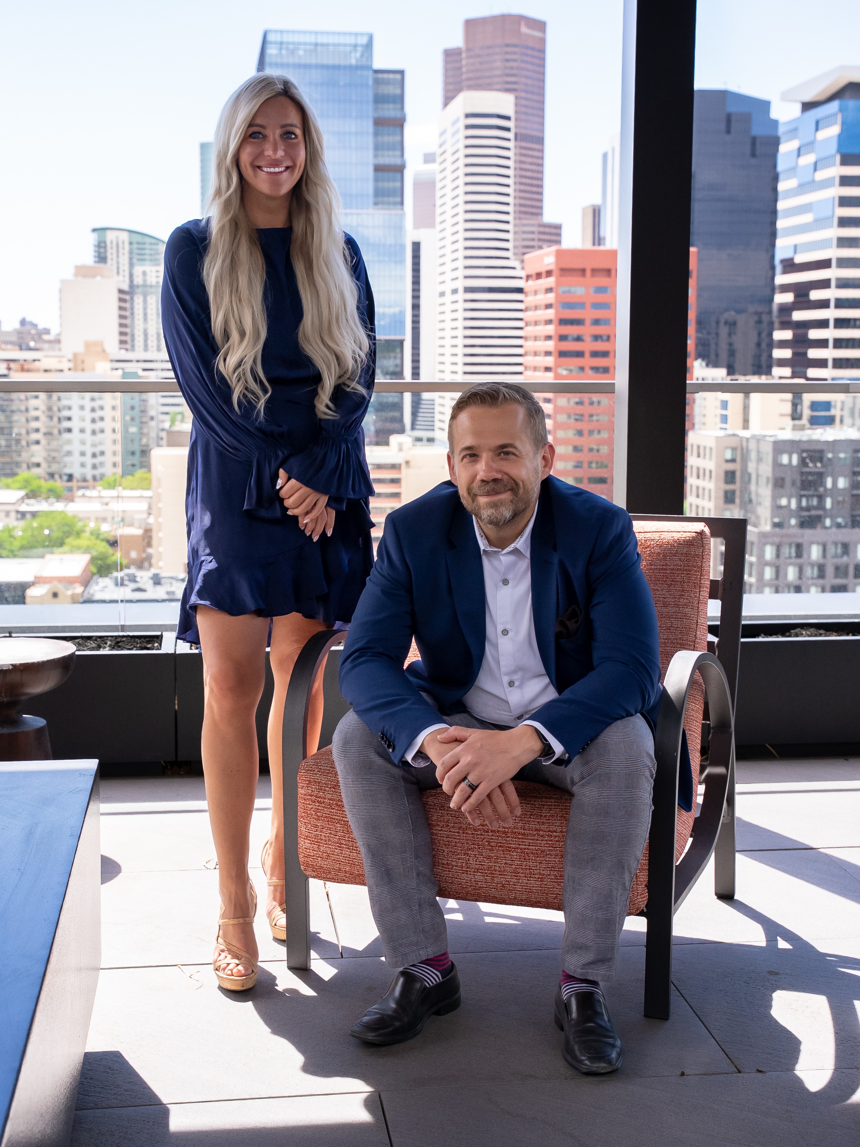 A woman and a man in business attire pose in a modern office with city skyline view behind them.