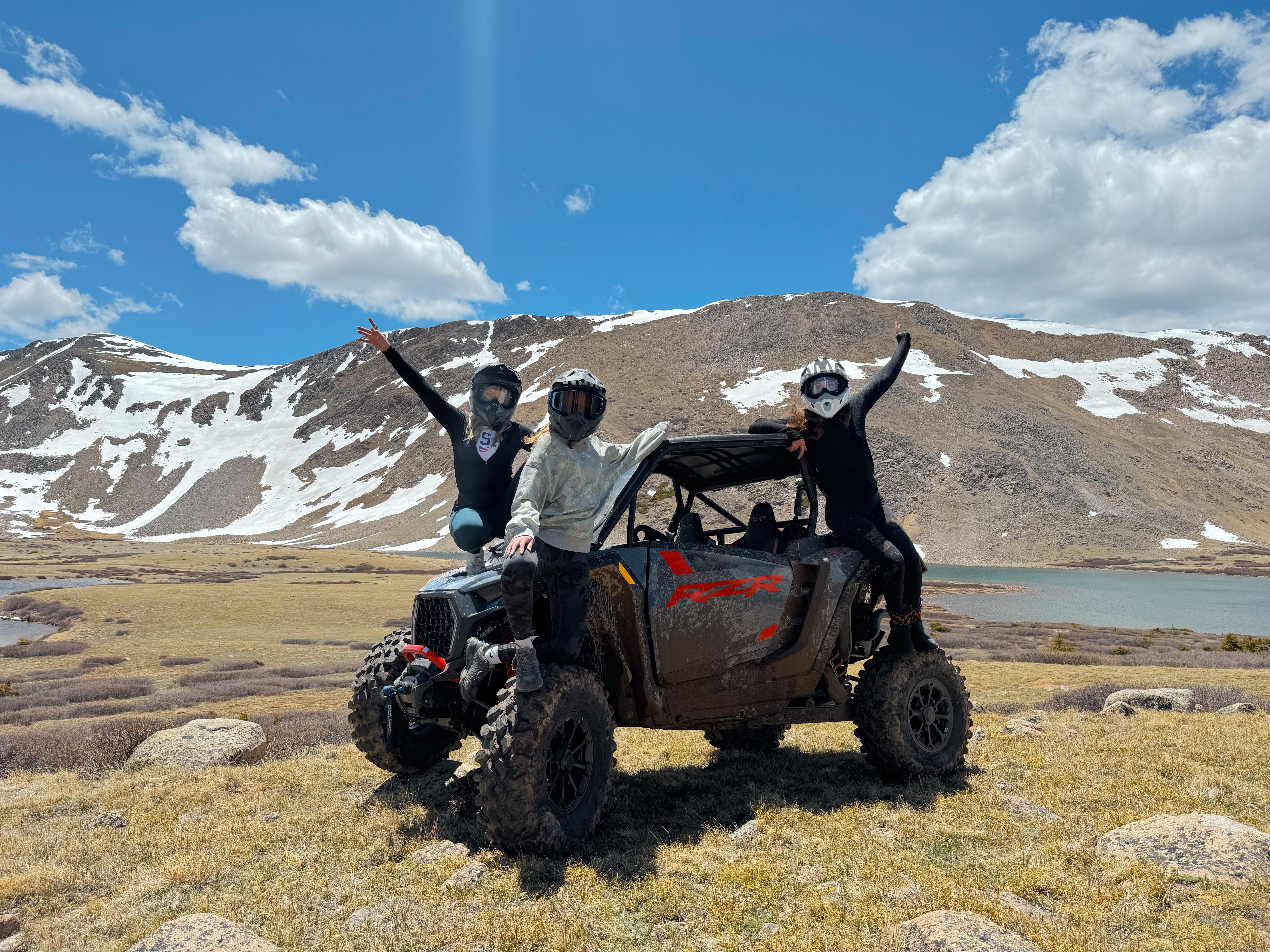 Three people standing next to a black off-road vehicle in a mountainous landscape with snow patches and a lake, under a partly cloudy sky.