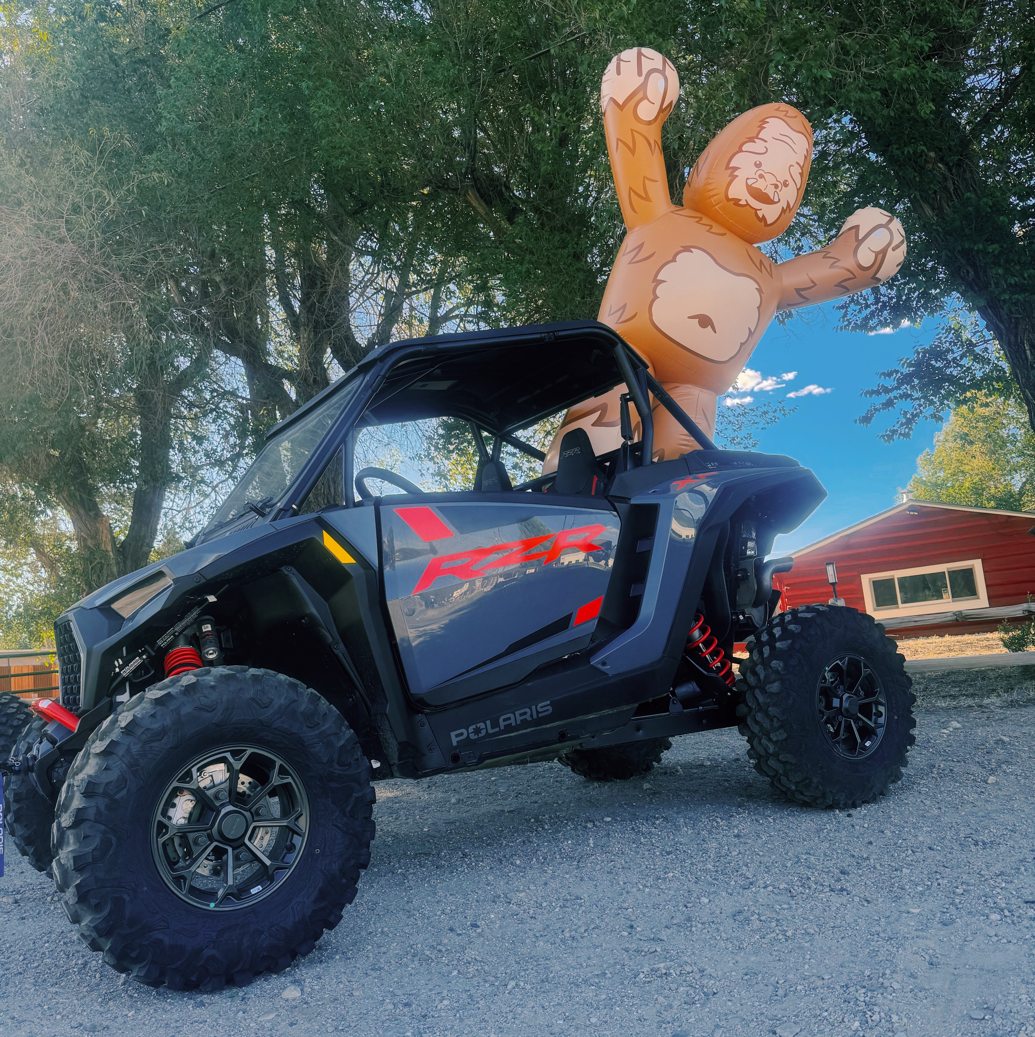 Polaris off-road vehicle parked outdoors with a large inflatable monkey in the background, trees and a building visible.