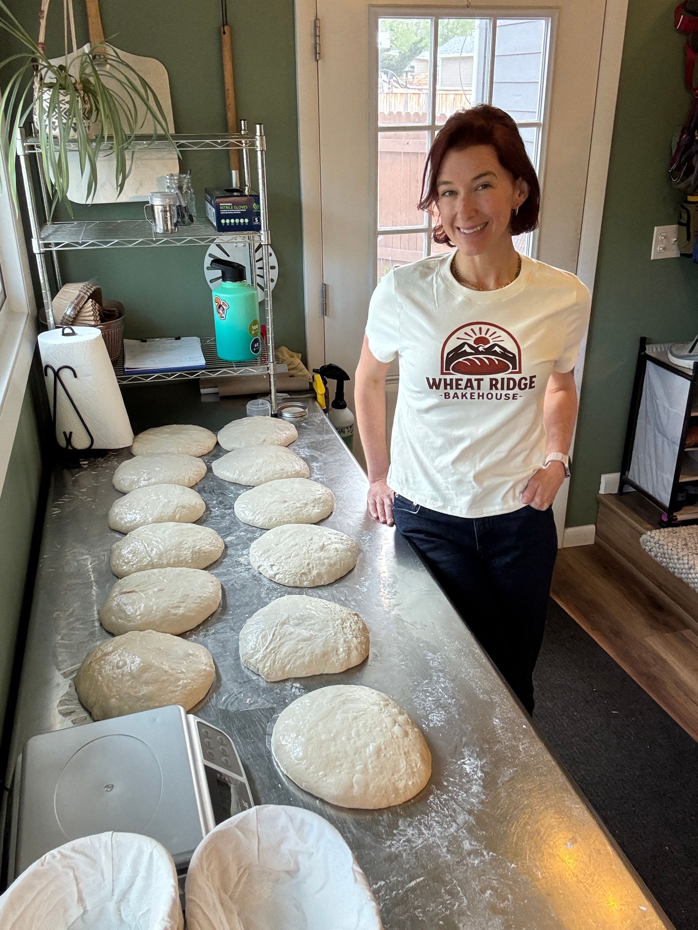 Woman standing in a kitchen with dough balls on a countertop, window in background, wearing a white T-shirt and dark pants.