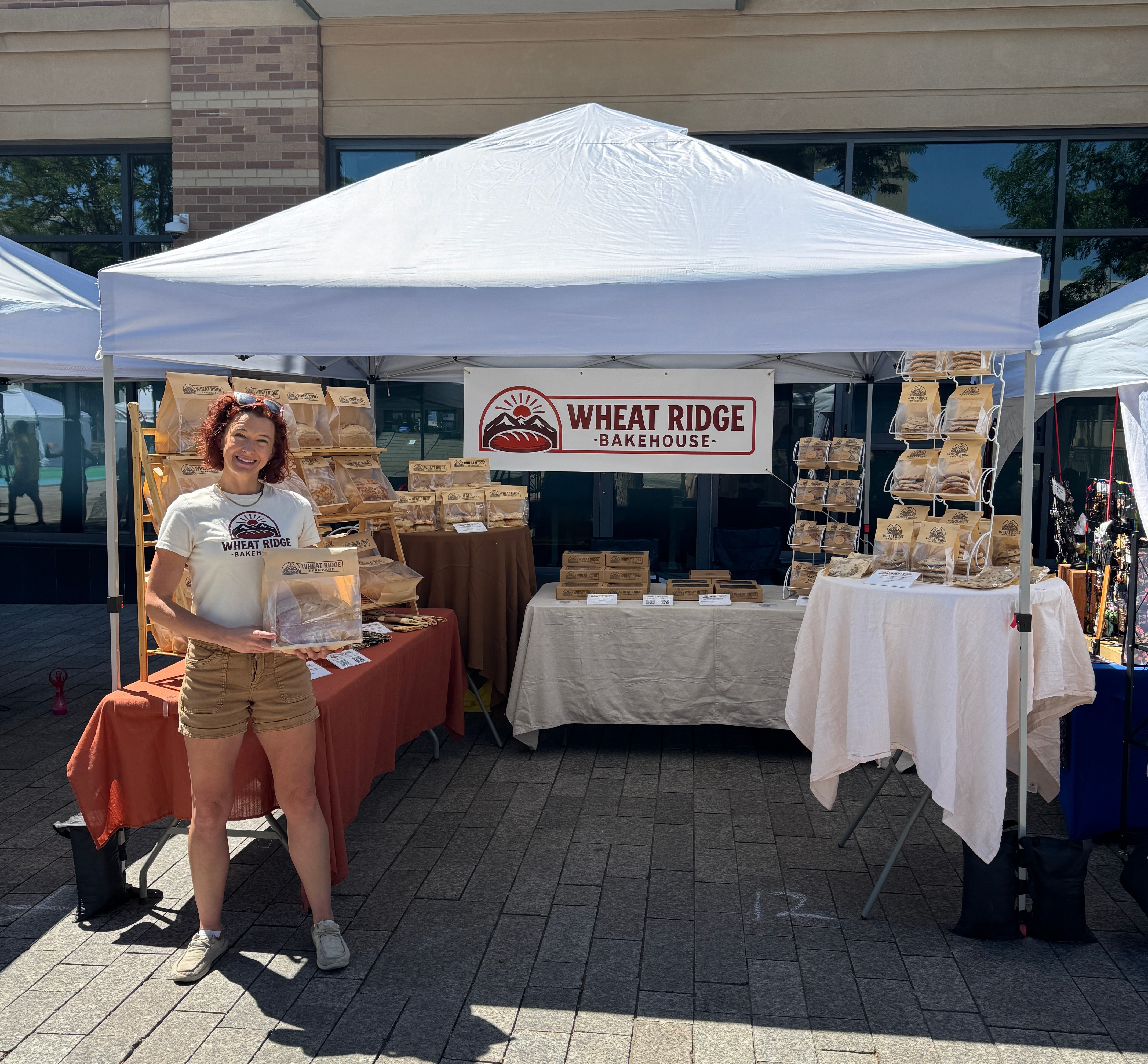 Woman standing in front of a bakery booth with bread displays, smiling, outdoors on a sunny day.