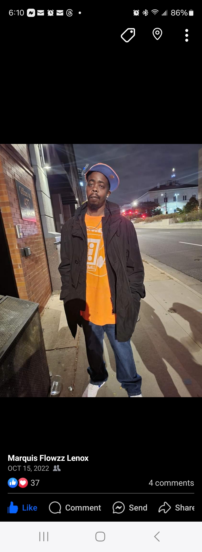 Person standing on sidewalk at night, wearing orange shirt, black jacket, and blue jeans, with streetlights and buildings in background.