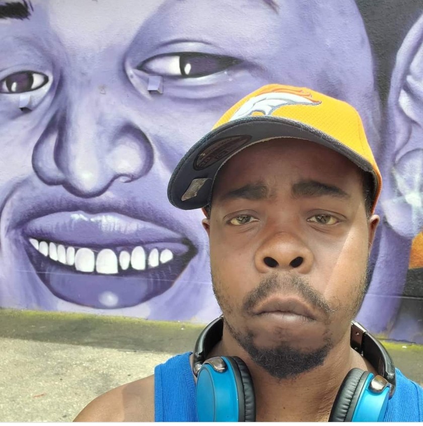 Young man wearing a yellow cap and blue headphones takes a selfie in front of a large purple mural of a smiling face.