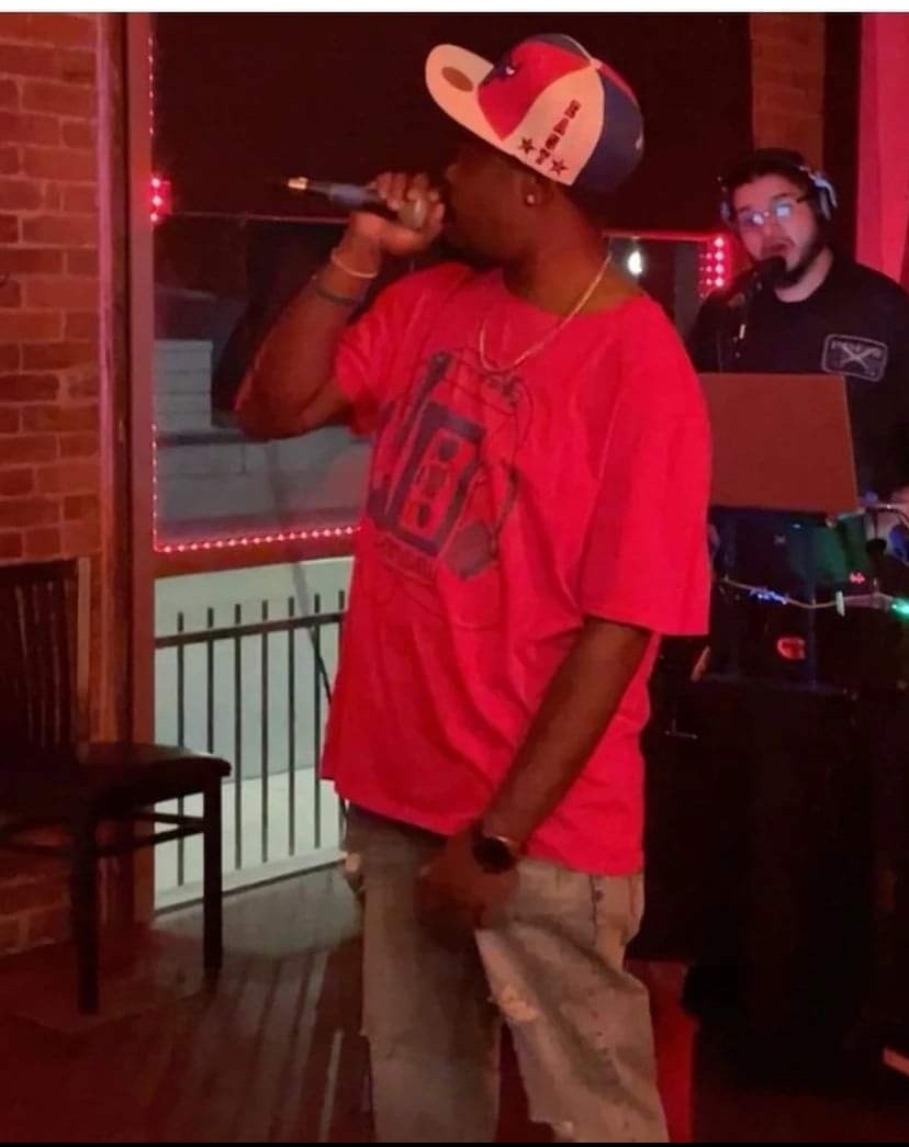 Person wearing a red shirt and a colorful cap, holding a microphone, with a DJ in the background, indoors at night.