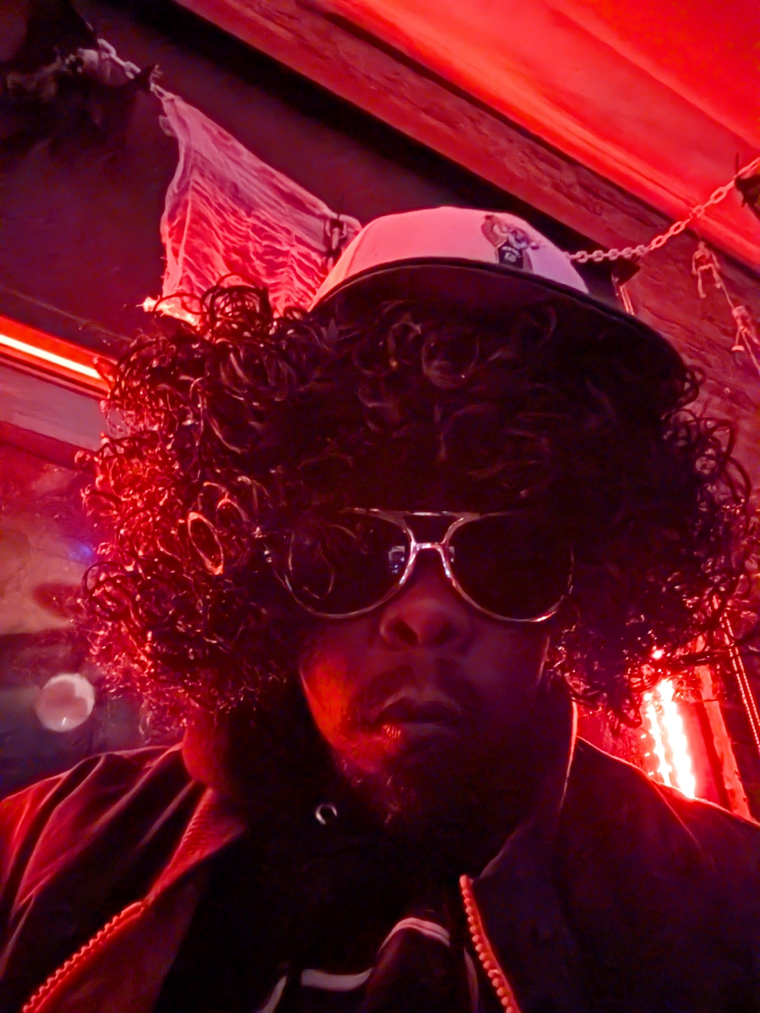 Person with curly hair wearing sunglasses and a hat, illuminated by red lighting, indoors.