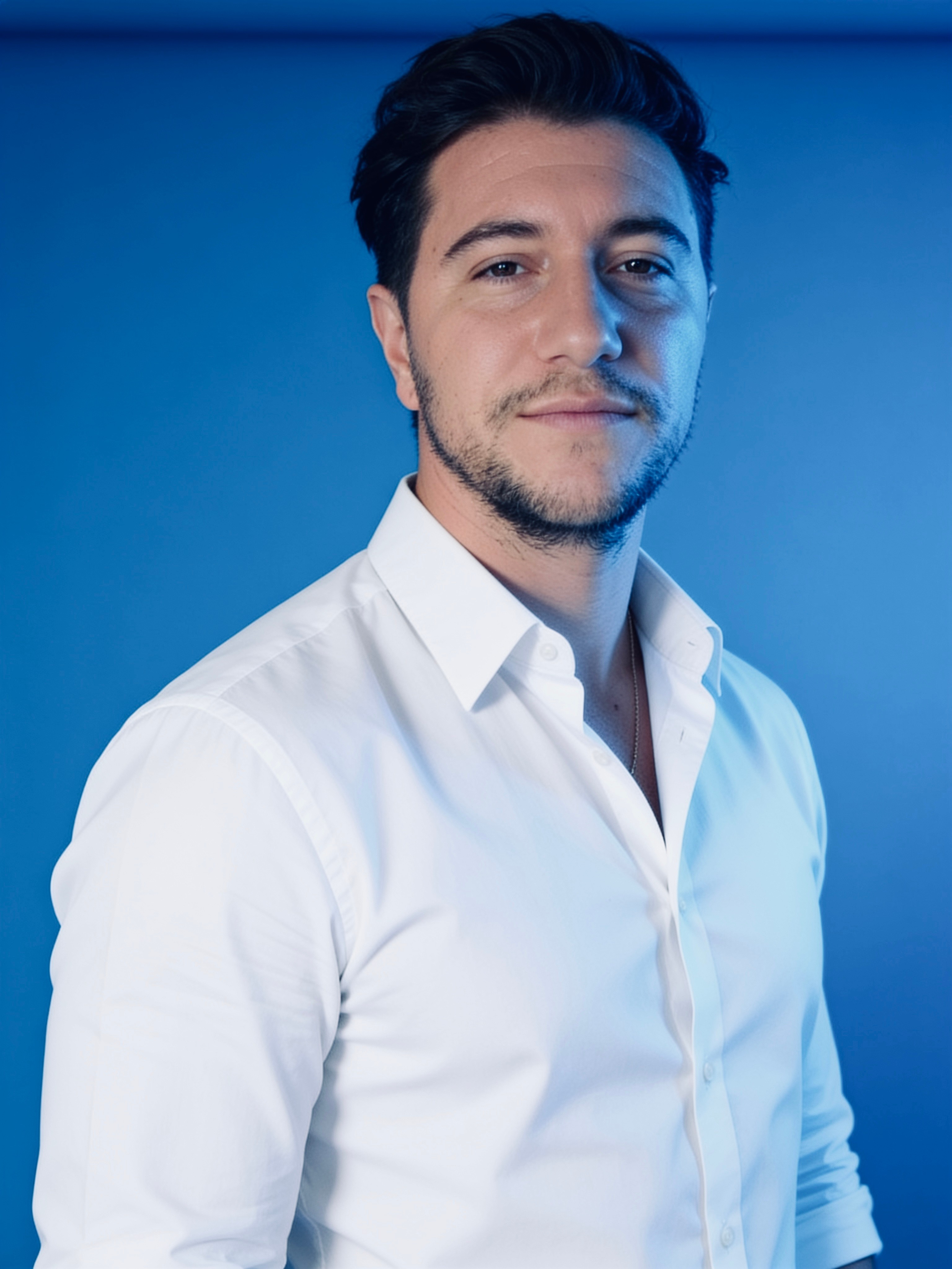Man with dark hair and beard wearing a white shirt, standing against a blue background.