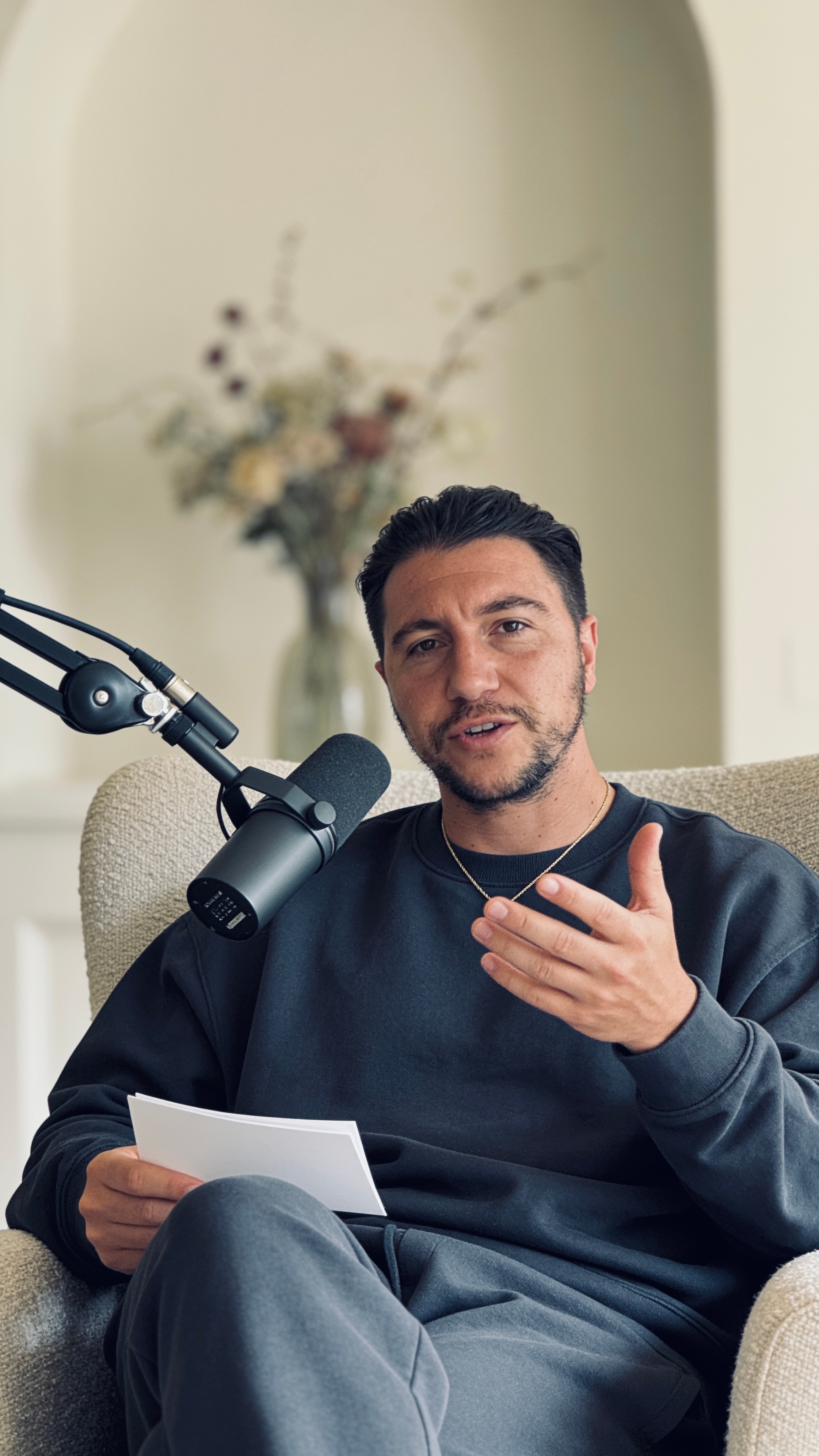 Man sitting on a beige armchair, speaking into a microphone, holding a paper, with a blurred floral background.