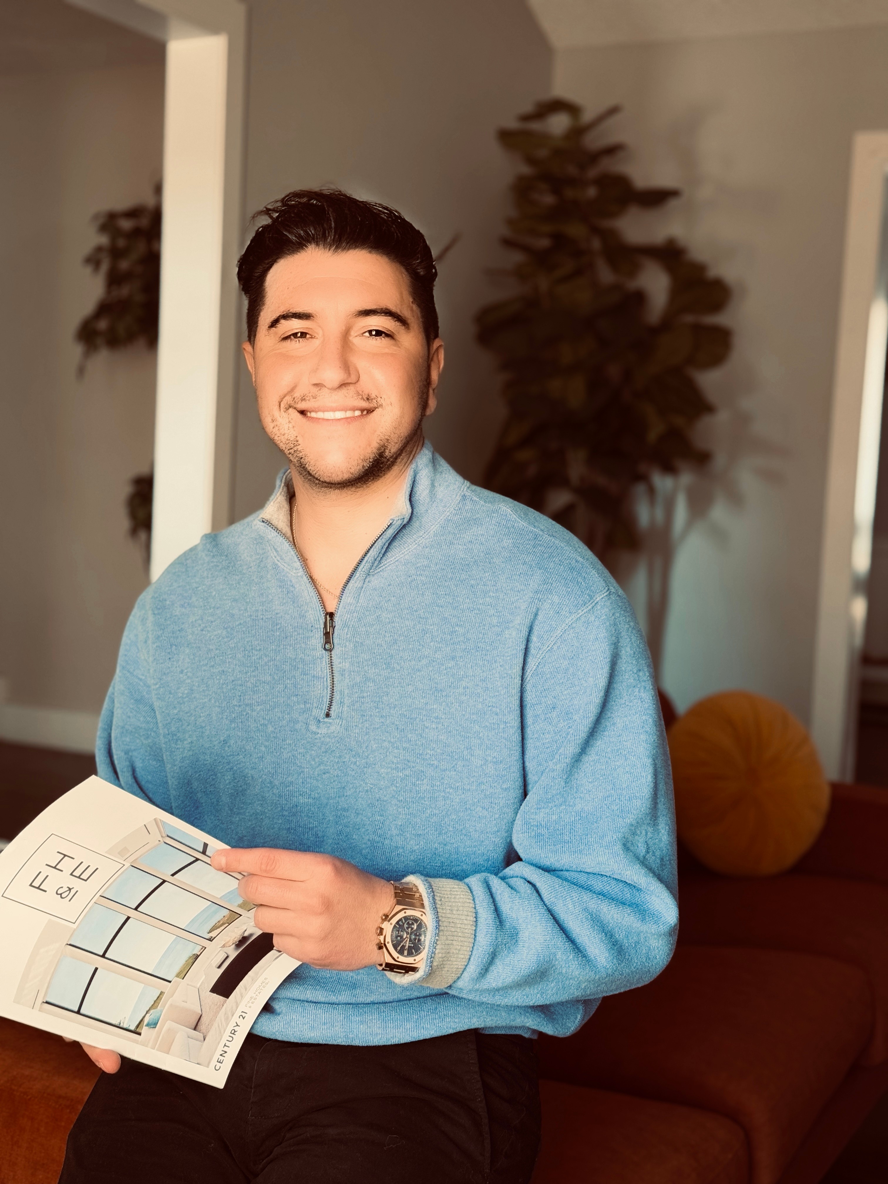 Smiling young man holding a magazine indoors, wearing a light blue sweater and a watch, with plants and furniture in background.